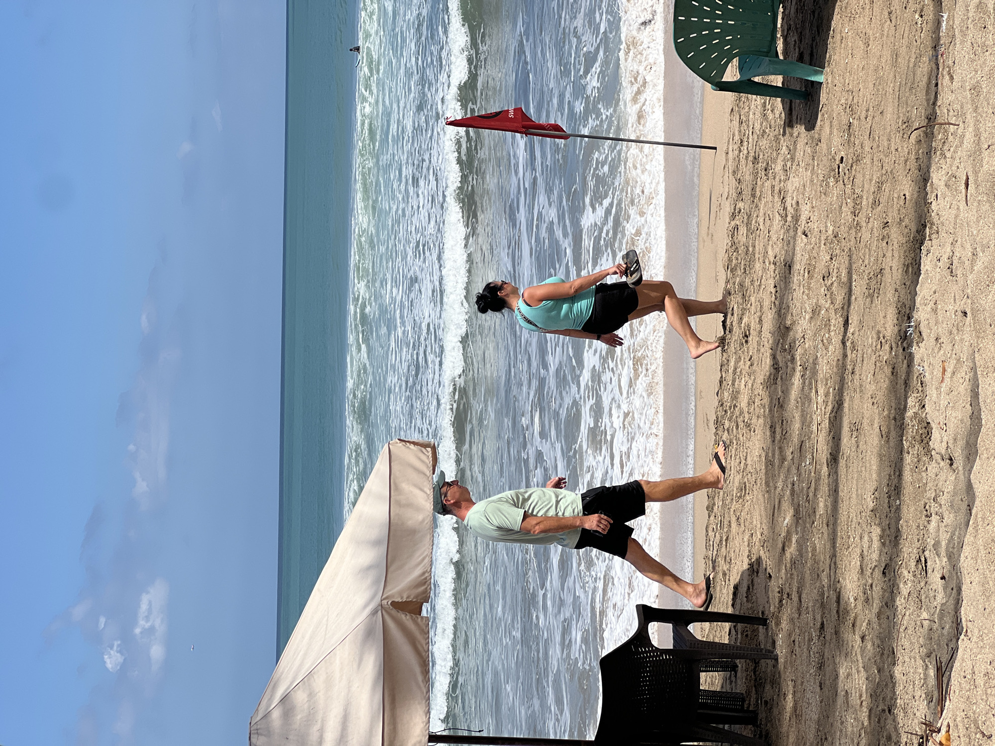 This image captures a sunny day at Kuta Beach, Indonesia. Two people, a man and a woman, are walking along the sandy shore. The man, wearing a green t-shirt and black shorts, walks towards the left of the frame, holding a phone in his right hand. The woman, in a teal tank top and black shorts, walks parallel to him, carrying a pair of flip-flops in her left hand.
In the background, the turquoise waters of the ocean stretch out to the horizon, with white waves breaking on the shore. A red flag, possibly indicating swimming conditions, stands planted in the sand. Further out in the water, a lone surfer can be seen on a surfboard. In the foreground, a beach umbrella casts a shadow over the sand, with black chairs positioned beneath it. A green plastic chair is visible to the right. The sky is a bright blue with scattered white clouds. An airplane can be seen in the distance.
