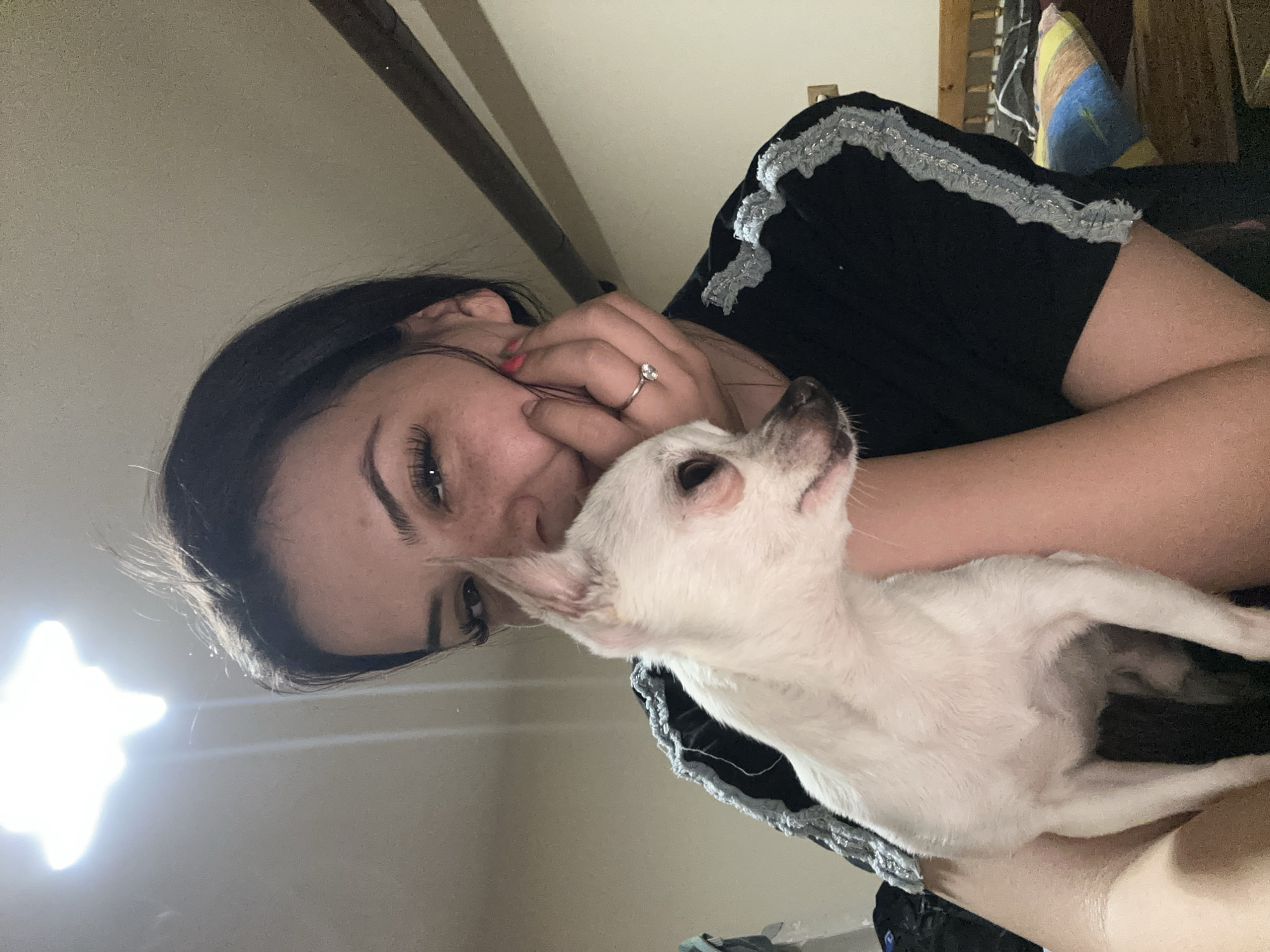 A light-skinned woman with dark hair, wearing a black top with silver trim, is holding a white Chihuahua-type dog. She is wearing a ring on her left hand. The environment appears to be indoors, possibly a bedroom, with a wooden beam visible in the background. The location is unknown.