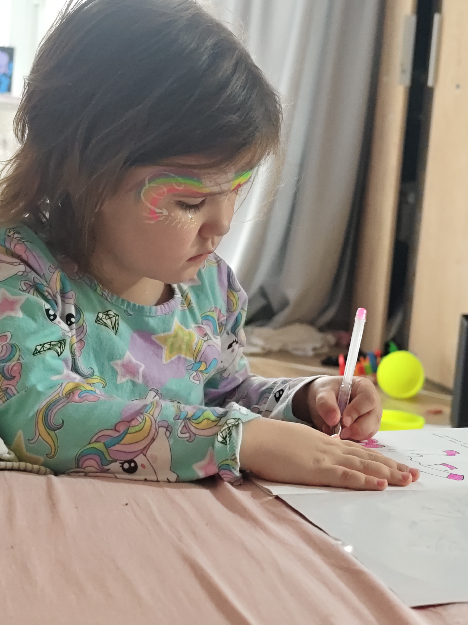 An image captured in Chaikovsky, Russia, depicts a young child seated indoors, engaged in a drawing activity. The child, possessing medium-brown hair, is shown in a partial profile view, looking downwards and to the right of the frame, concentrating on the task. Face paint is applied to the child's left side of the face (viewer's right), featuring a multi-colored rainbow arc, white swirling patterns, and glitter positioned beneath the eye. The child is dressed in a long-sleeved, light turquoise shirt adorned with numerous multicolored unicorn and star patterns. In their right hand, the child holds a clear pen with a pink cap, actively coloring or drawing on a white sheet of paper. The paper exhibits outlines resembling paper doll clothing items, with portions already filled in with pink. The child's arms rest upon a wrinkled pink fabric surface, consistent with a blanket or duvet. The background on the left consists of a blurry light-colored wall. To the right, a light-toned curtain partially obscures a light wooden door or cabinet, along with a dark vertical frame. On the floor in the background, a bright yellow spherical object and other indistinct colorful items are visible.