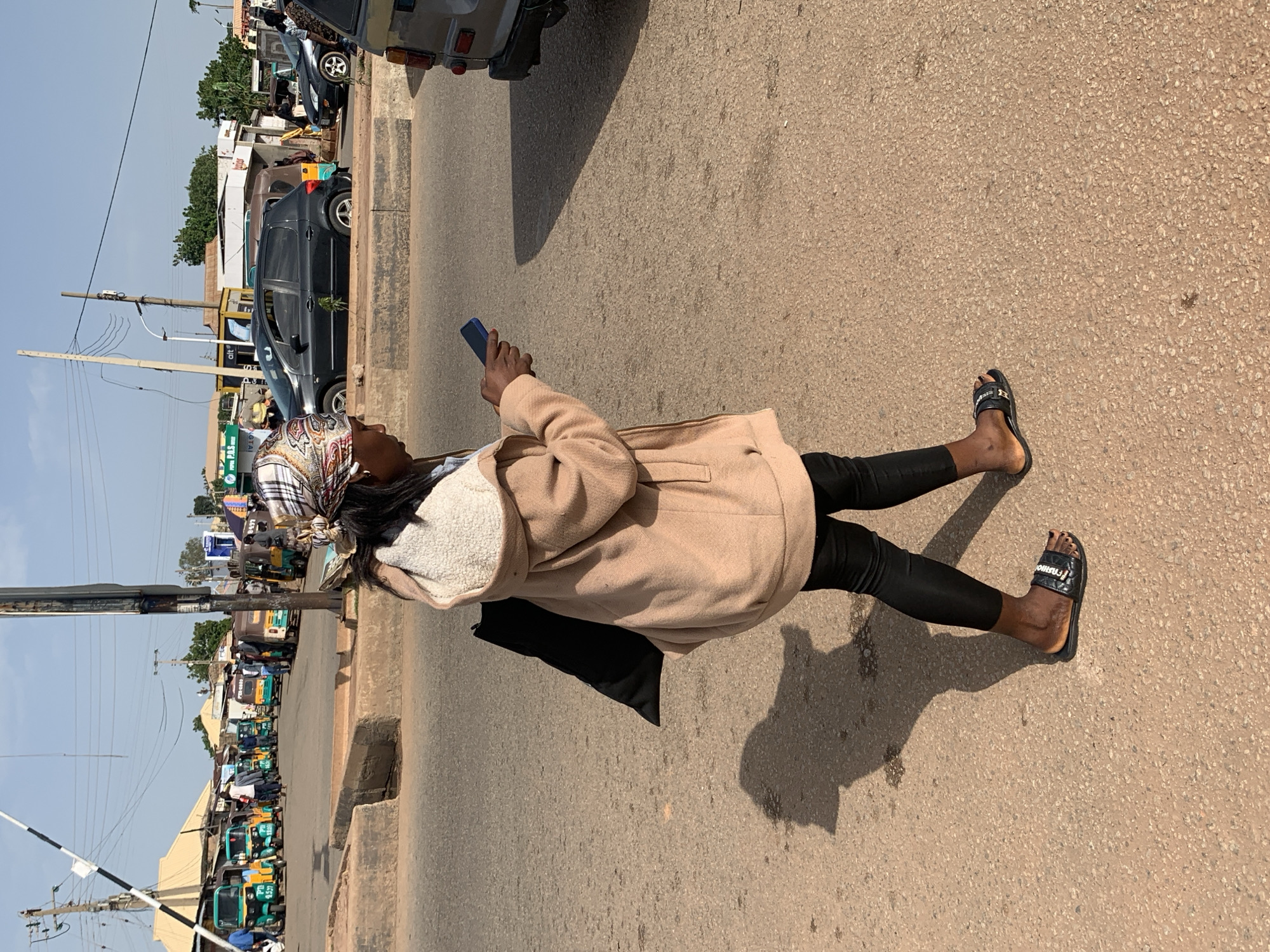 A medium shot captures a woman with dark skin from the side, walking on a paved road in Gura Top, Nigeria. She is wearing a beige hooded coat with a sherpa lining, black leggings, and black slides with a diamond-patterned strap. A black bag hangs from her left shoulder. Her head is covered with a patterned headscarf, and she is looking down at a blue mobile phone in her right hand. In the background, a variety of vehicles, including numerous green and yellow motorized rickshaws and several cars, are parked or moving on the road. Buildings and utility poles are visible further in the distance. The sun is casting a shadow of the woman on the ground to her left.