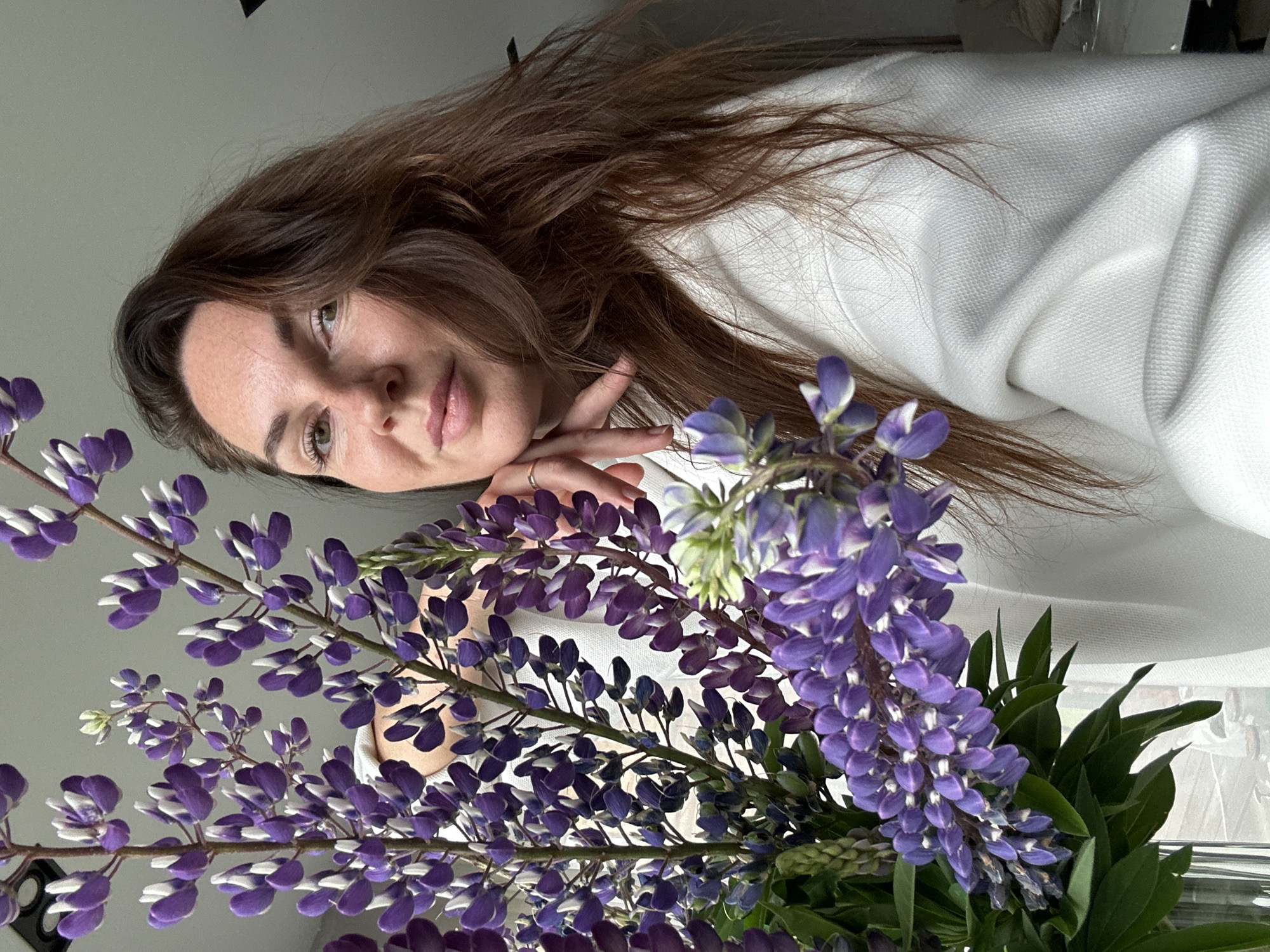 A woman with brown hair and green eyes is pictured in a close-up shot, holding a bouquet of purple lupine flowers. She is wearing a white sweater and her left hand is resting on her chin. The background is neutral, with a gray wall and a hint of a window on the left. The image appears to be taken indoors, likely in a residential setting. The woman is looking towards the camera with a slight smile. The lupine flowers are in full bloom and form a vibrant contrast to the woman's white attire. The scene is illuminated by natural light.