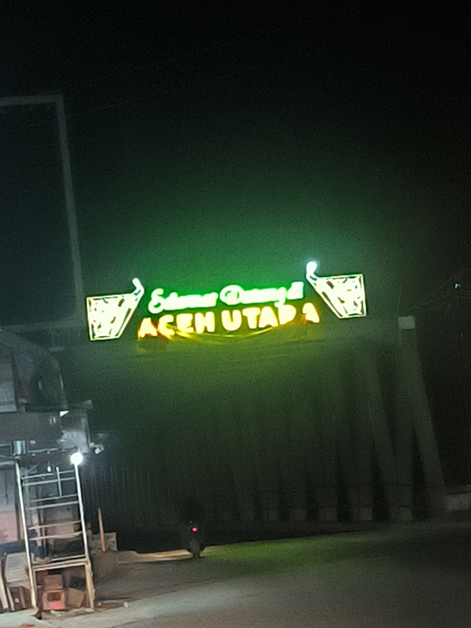 At night, the image depicts a roadside establishment in Kotapantonlabu, Indonesia, displaying a neon sign that reads "Selamat Datang di ACEH UTARA." The sign is illuminated in green and yellow, featuring stylized images of iced tea glasses on either side of the text. A simple metal structure, possibly a small kiosk or food stall, is visible on the left, with some visible contents. The surrounding environment suggests an urban or semi-urban setting.