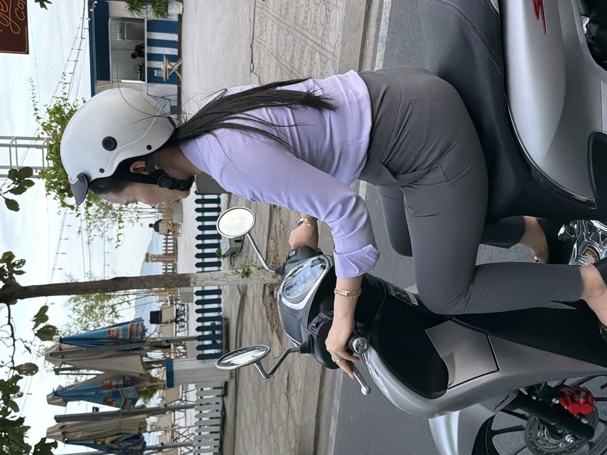A woman wearing a white helmet, a light purple collared shirt, and grey pants is riding a silver scooter in Nha Trang, Vietnam. She is looking to her left. The scooter's handlebars and front panel are visible. In the background, there are several closed umbrellas, a white picket fence, and a building with blue and white stripes. The sky is overcast.