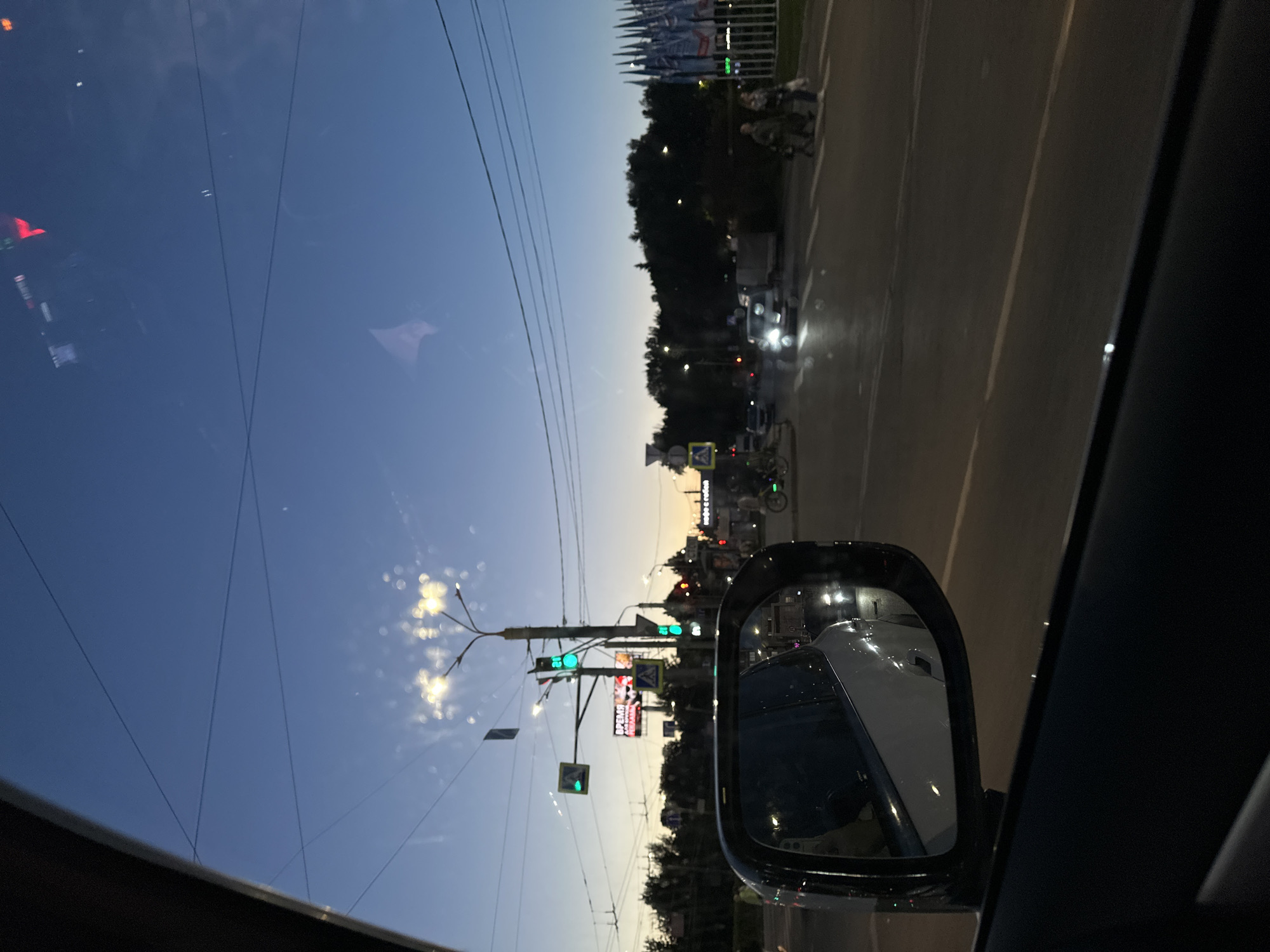 The image shows a view from inside a car at dusk, likely in Nizhny Novgorod, Russia. The scene is a street with a traffic light, trees, and buildings in the background. A side-view mirror of the car reflects the scene. Traffic lights are visible, with some showing green signals. There are also streetlights and some lights from buildings in the distance. A few people are visible walking on a crosswalk. The sky is a clear blue, transitioning to a lighter hue near the horizon, suggesting either sunrise or sunset. The overall mood is calm and serene, typical of early evening. There are no discernible emotional cues from the subjects.