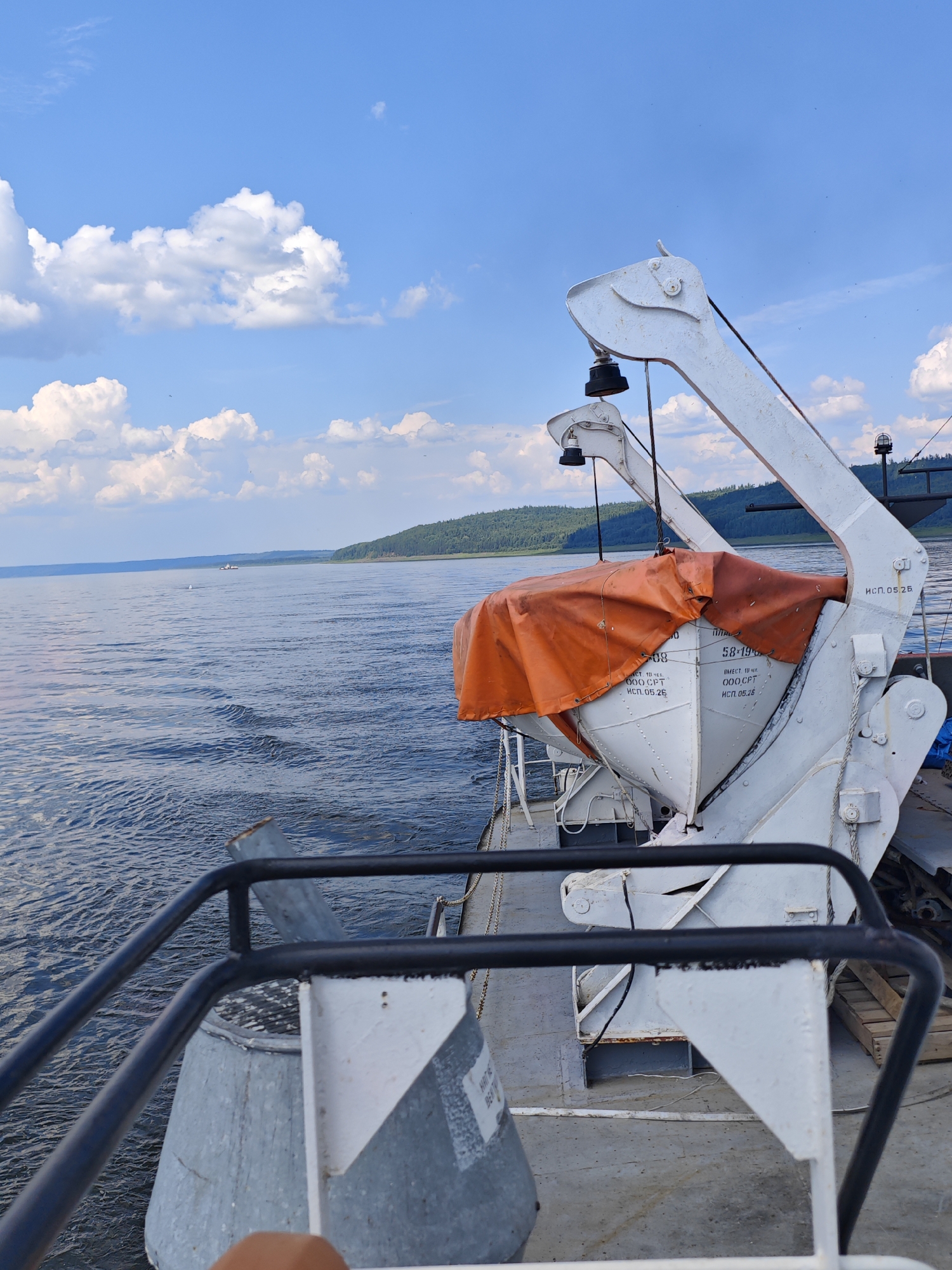 The image depicts a view from the deck of a boat on a wide river under a partly cloudy blue sky. In the foreground, a white lifeboat, covered by an orange tarp, is secured by a davit crane system. The crane is white and appears to be made of metal. Part of the boat's railing, a black metal bar, is visible in the lower left quadrant of the image.
The river is calm with gentle ripples reflecting the sky. In the distance, a forested, hilly shoreline stretches across the horizon. A small vessel can be seen in the middle ground on the river. The overall scene suggests a serene day on the water, likely during daytime.
While the image does not explicitly show the city of Podkamennaya Tunguska, Russia, the described elements of a wide river, forested shores, and riverine transport are consistent with the geographical characteristics of the region.