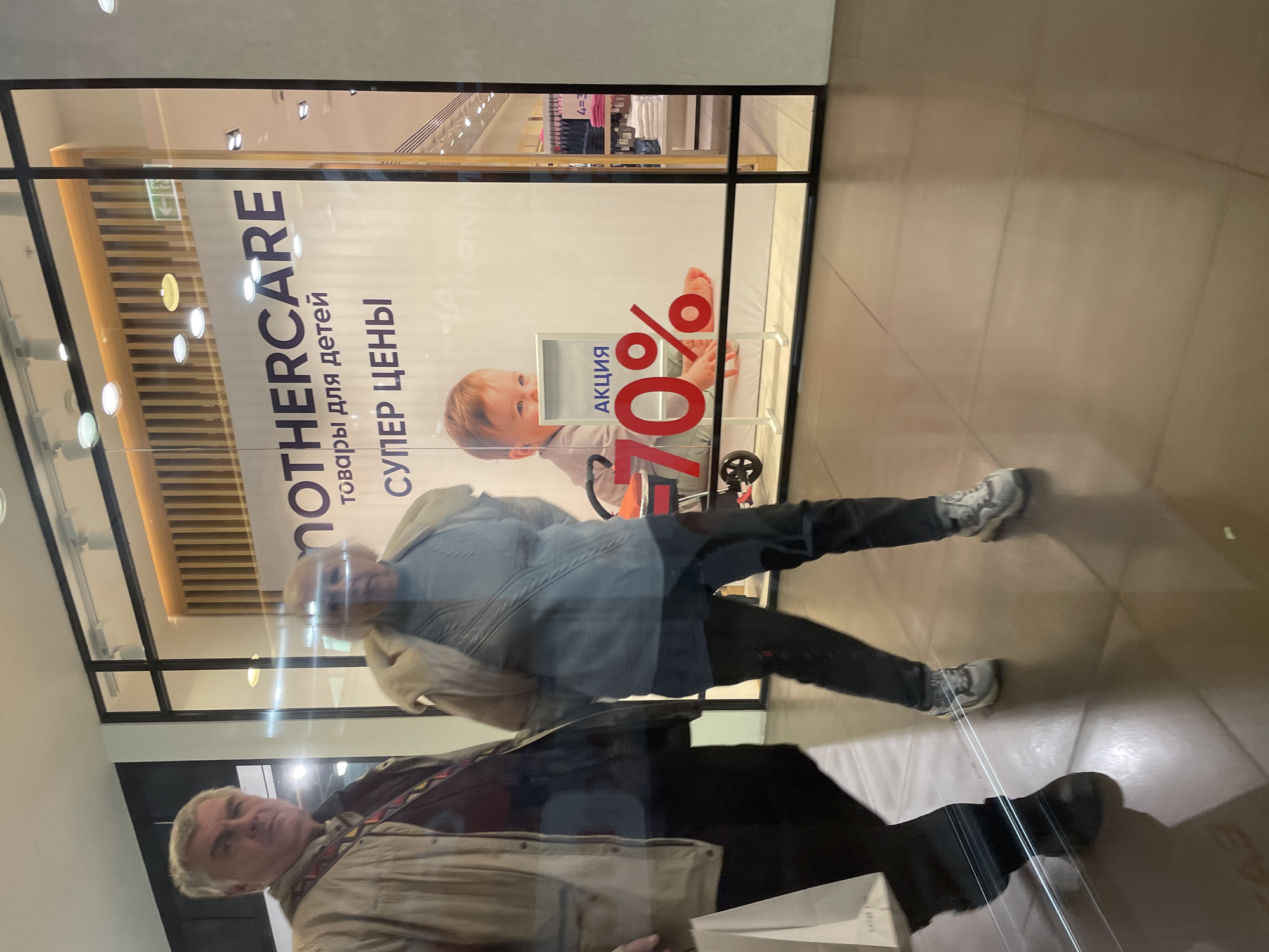 The image shows a reflection of two individuals in a shop window. The store is identified by a sign for "MOTHERCARE" with text in Russian ("товары для детей" - goods for children; "СУПЕР ЦЕНЫ" - super prices; "АКЦИЯ - 70%" - promotion - 70%).
The taller individual is wearing dark pants, a light-colored hoodie with a jacket over it, and sneakers. The second individual wears dark trousers, a brown shirt, and a beige jacket.
Based on the presence of the Russian language and the "MOTHERCARE" store, it is likely that this photograph was taken in Saint Petersburg, Russia, though the exact location is not discernible from the image. The individuals are reflected in the store window while seemingly walking past the storefront. The photograph has a rotated view.
