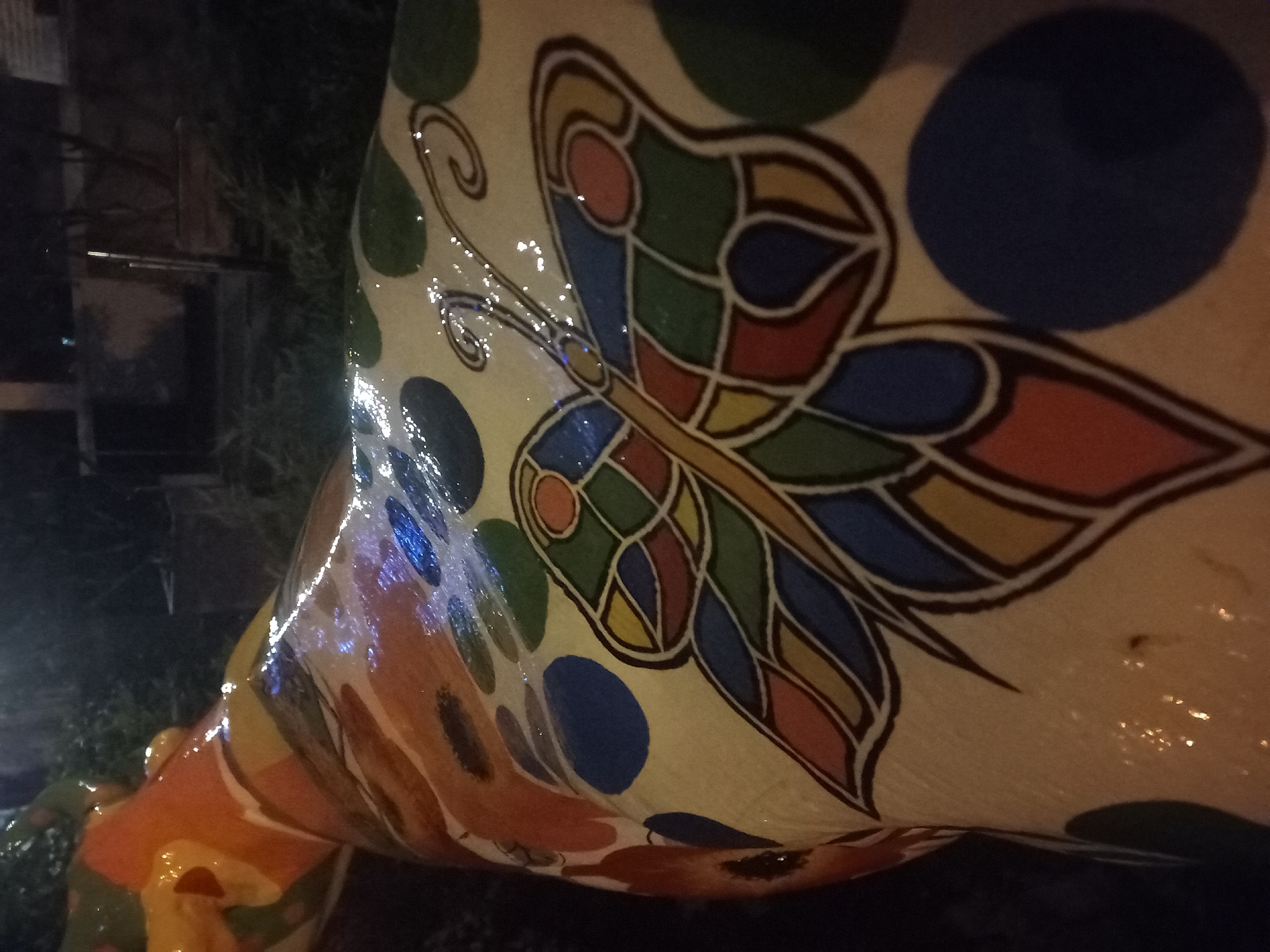 A close-up shows a portion of a public art object, specifically a painted statue, likely of an animal (e.g., a cow, horse), in Kyiv, Ukraine. The statue's surface is decorated with a colorful butterfly design, featuring geometric shapes in shades of blue, green, red, yellow, and orange, outlined with a dark brown or black color. Randomly placed, there are dark blue and green circles around the butterfly. The base color of the statue is off-white. The surface is glossy and reflects light. The background is dark and indistinct, suggesting the photo was taken at night or in low light. Some vegetation is visible in the background, along with hints of urban elements, like a window. The statue is positioned outdoors, most likely in a public space within the city.