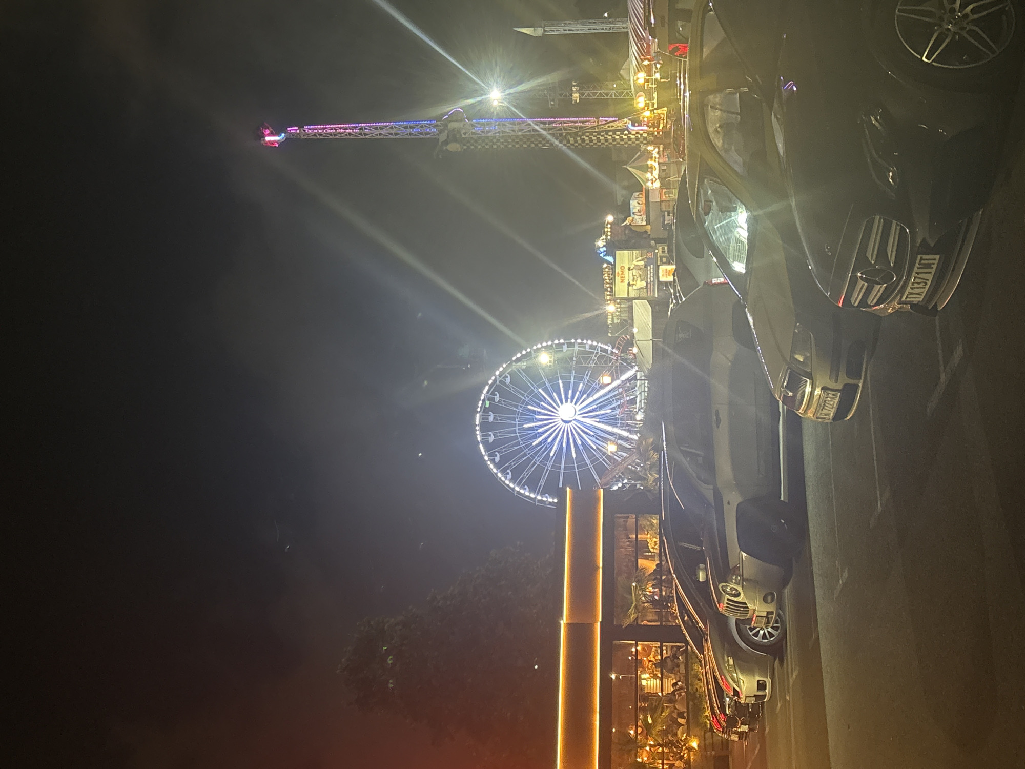 This is a nighttime photograph depicting a vibrant amusement park or fairground scene. In the background, a towering drop tower ride with colorful pink, purple, and blue lights extends high into the dark sky, with a person visible at its apex. To the left of the frame, a large Ferris wheel glows brightly with white and blue lights, illuminating the surroundings. Numerous bright lights from the rides and stalls create prominent lens flares across the dark sky.
In the mid-ground and foreground, various structures, likely stalls or game booths, are lit with warm yellow and orange lights. A tall, cylindrical pole, also glowing yellow-orange, stands prominently towards the right of the Ferris wheel. To the right side of the image, several cars are parked, indicating visitors to the event. A lighter-colored SUV in the foreground has a visible license plate that reads "BN72 ZNA." Behind it, a darker car, possibly a Mercedes-Benz, shows the license plate "DL67 JNT." On a sign in the middle ground, the word "NEMO" is partially visible. The overall atmosphere is lively and bright, characteristic of a nocturnal entertainment venue.