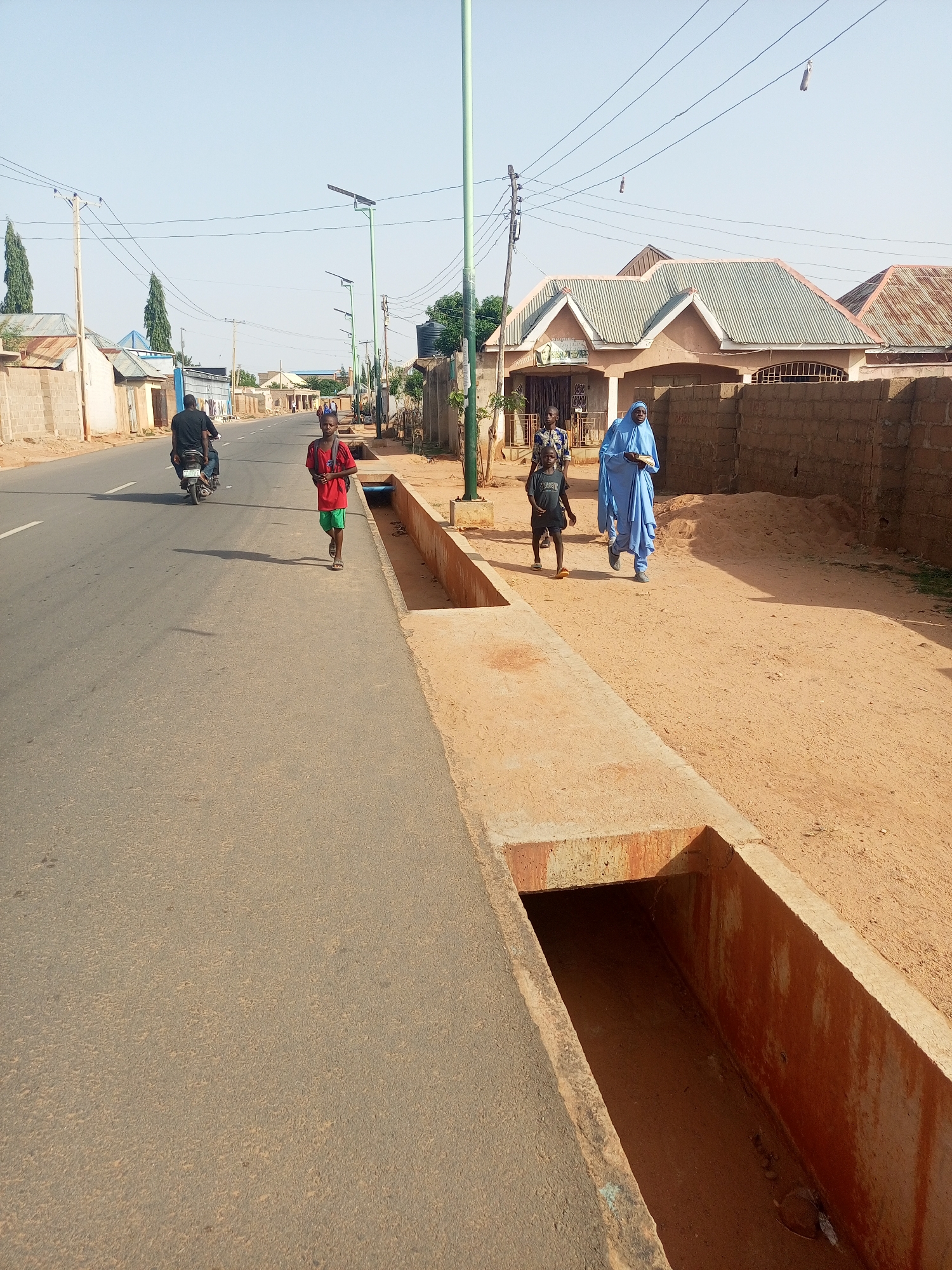 The image shows a street scene in Tempure, Nigeria. A paved road runs along the left side of the frame. A motorcycle with a rider is on the road. A young boy in a red shirt and green shorts walks along the edge of the road. To the right, a dirt path runs parallel to the road, separated by a concrete drainage ditch. On this path, an adult woman in a light blue hijab walks with two younger children. The backdrop consists of various single-story buildings and houses, with power lines overhead. The sky is clear and light blue. Street lamps line the street.