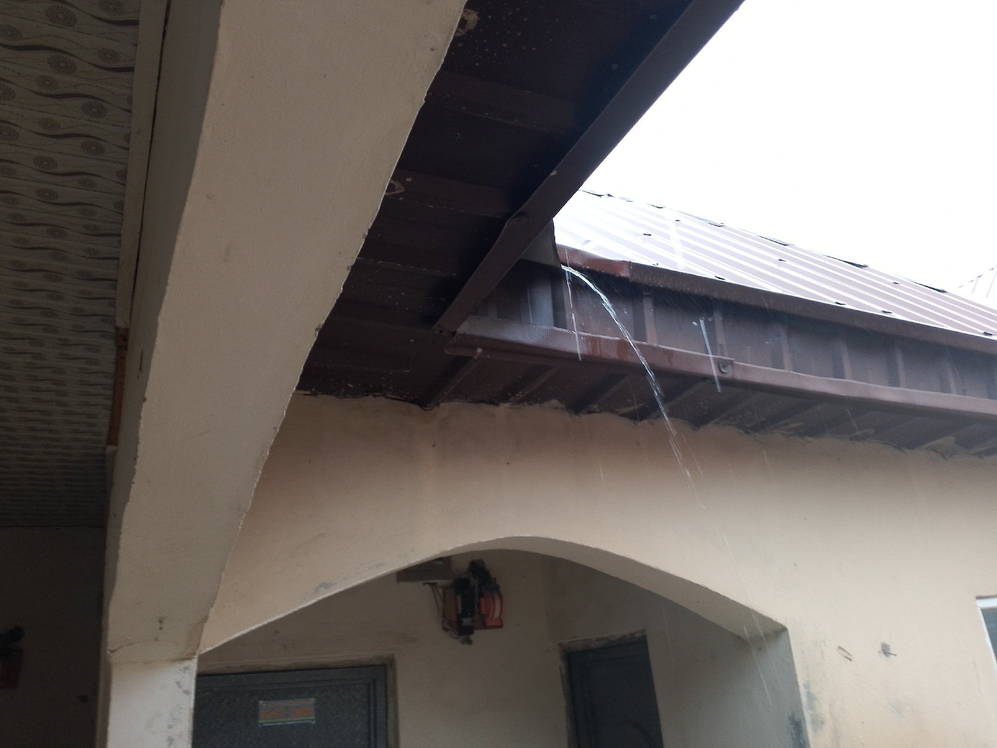 The image depicts an outdoor scene under a light sky, likely during a rain shower. A dark brown, corrugated metal roof overhangs a light-colored wall. Water is visibly streaming down from the edge of the roof. Below the overhang, an arched opening reveals a light beige wall with two dark doorways visible. A red and black electrical device is mounted on the wall within the arched opening. The ceiling of the covered area, to the left of the image, is patterned with a repeating circular motif. The overall impression is of an exterior area of a building experiencing rainfall. The location is specified as Unknown, Unknown.
