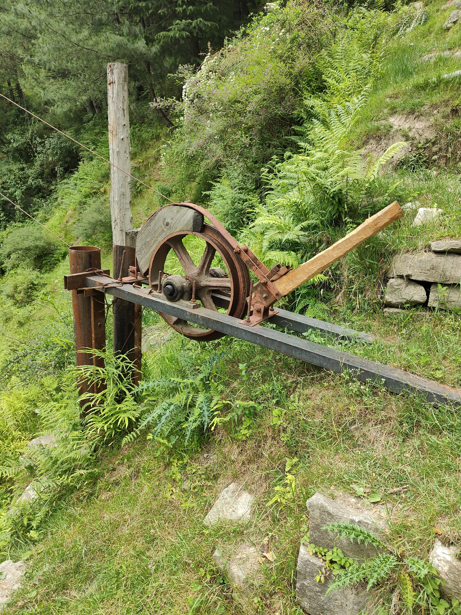 An old, rusty winch mechanism is situated on a grassy slope in Vashisht, India. The winch features a large wooden wheel with a metal rim, connected to a metal frame and a wooden lever. It is positioned next to a weathered wooden post and a metal pole. The surrounding environment is lush with green vegetation, including ferns and trees. The ground is covered in grass and scattered rocks, with a stone wall visible in the background. There are no people or animals present in the scene.