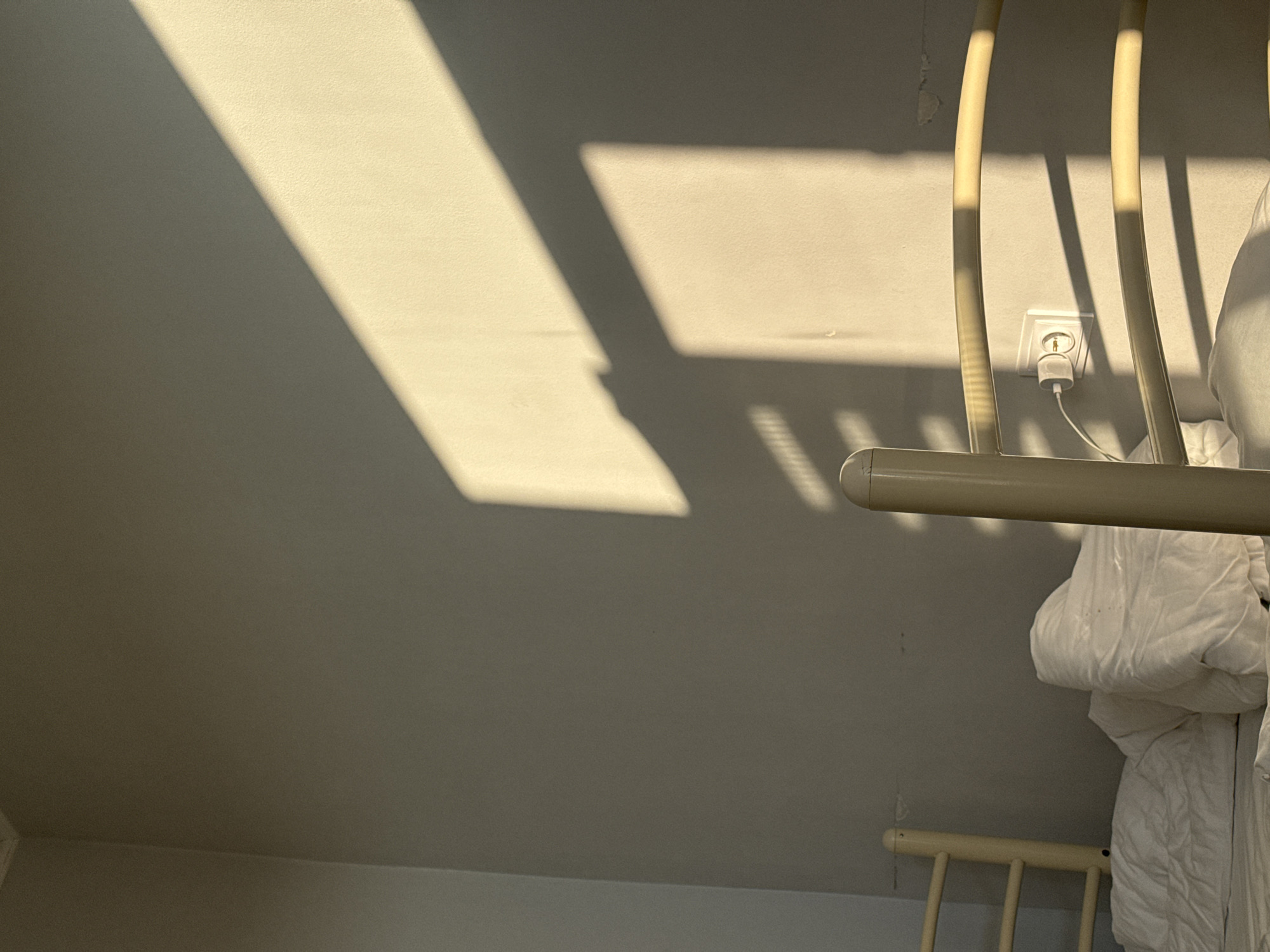 The image depicts a close-up view of a bed in a room, with sunlight casting strong shadows across a pale wall. In the foreground, a portion of a light-colored bed frame is visible, along with white bedding. A white electrical outlet with a connected charger is mounted on the wall to the right of the bed frame. The wall itself appears to be painted a neutral color and shows some minor imperfections. The lighting suggests it is either early morning or late afternoon, with the sun's rays entering from the side. The location of Chemerivtsi, Ukraine is mentioned, but there are no specific visual cues within the image to confirm this location. There are no people visible in the image, and no actions or interactions are taking place.