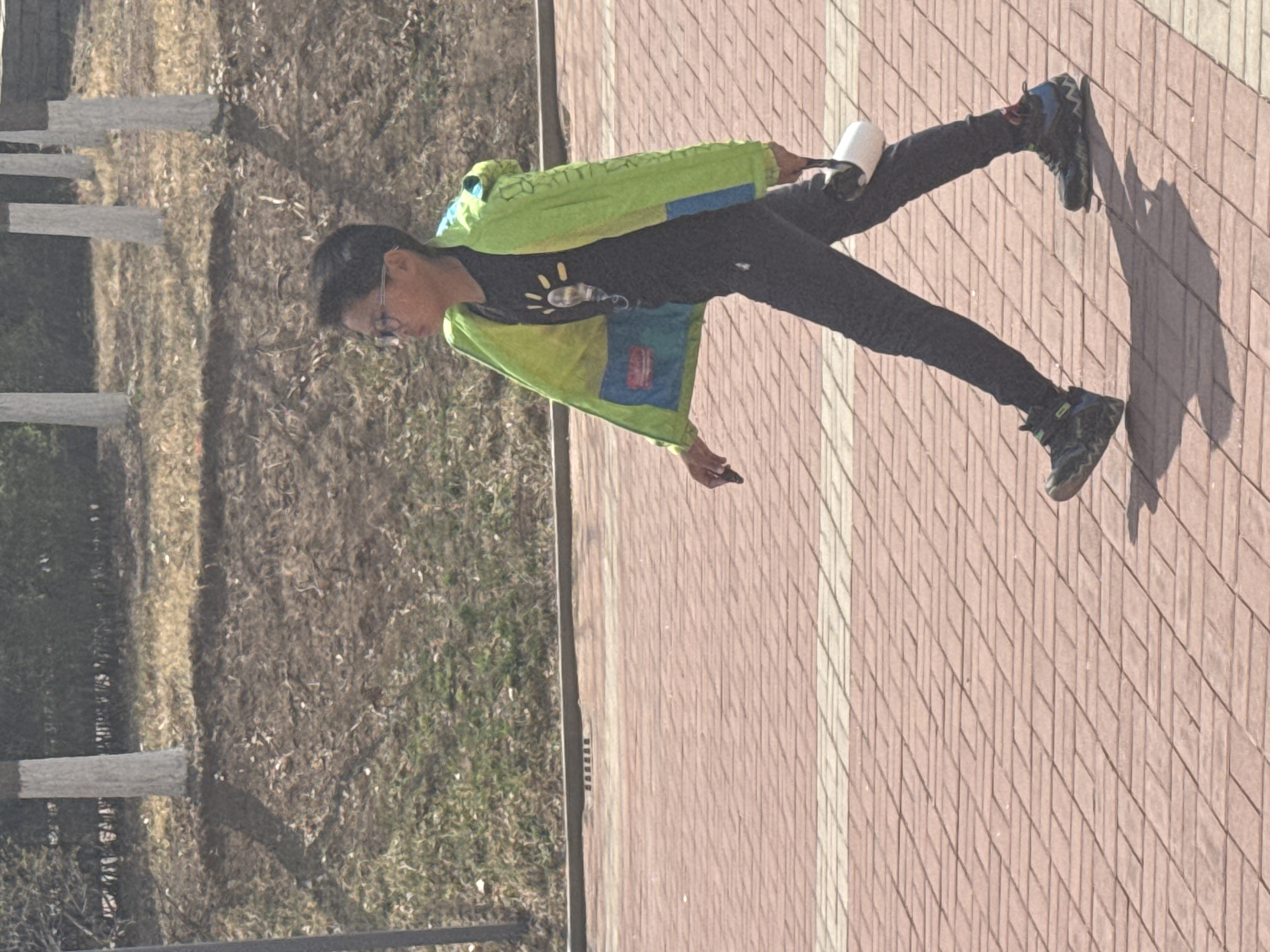 A person wearing a neon green jacket, black pants, and sneakers stands on a brick walkway next to an area of brown earth and vegetation. The person is holding a white cup. The location is likely Zhongtun, China, as indicated by the user.