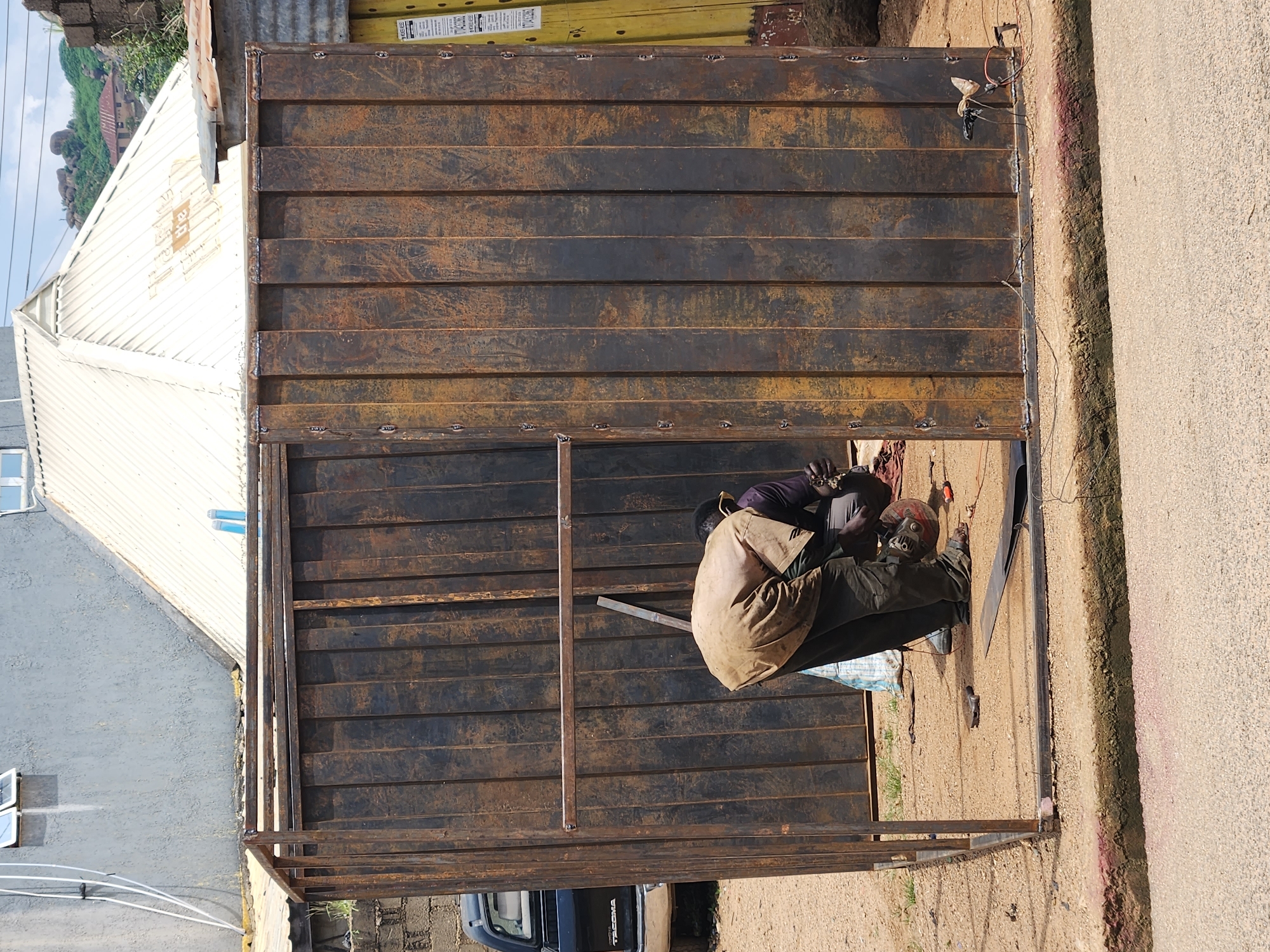 In Kabong, Nigeria, a man is seen working on a large, rusted metal structure, possibly a shipping container conversion or a makeshift stall. He is bent over, using a grinding tool on the metal. The structure is incomplete, with exposed framework and large, vertical metal panels that show signs of rust and wear. A black pickup truck is parked to the left of the structure, and in the background, parts of buildings with corrugated metal roofs and a grey textured wall are visible. The ground is dusty and uneven, with some loose tools and debris scattered around. The scene appears to be an outdoor workshop or construction site in an urban or semi-urban environment.