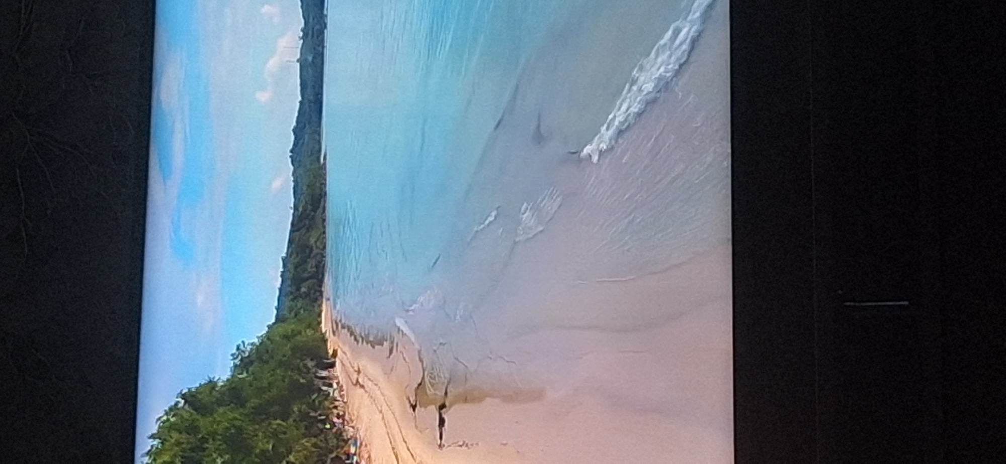 The image depicts a serene beach scene. The foreground is dominated by a sandy beach with gentle waves lapping at the shore. The water is a clear, tranquil turquoise, transitioning to a deeper blue further out. A line of lush green trees borders the beach, suggesting a tropical or subtropical environment. The sky above is a bright blue with scattered white clouds, indicating a sunny day. In the background, a rolling green landscape stretches towards the horizon, with hints of man-made structures like what appear to be communication towers in the distance. The overall impression is one of peace and natural beauty. The image does not provide any information that would indicate a location in Bila Tserkva, Ukraine.