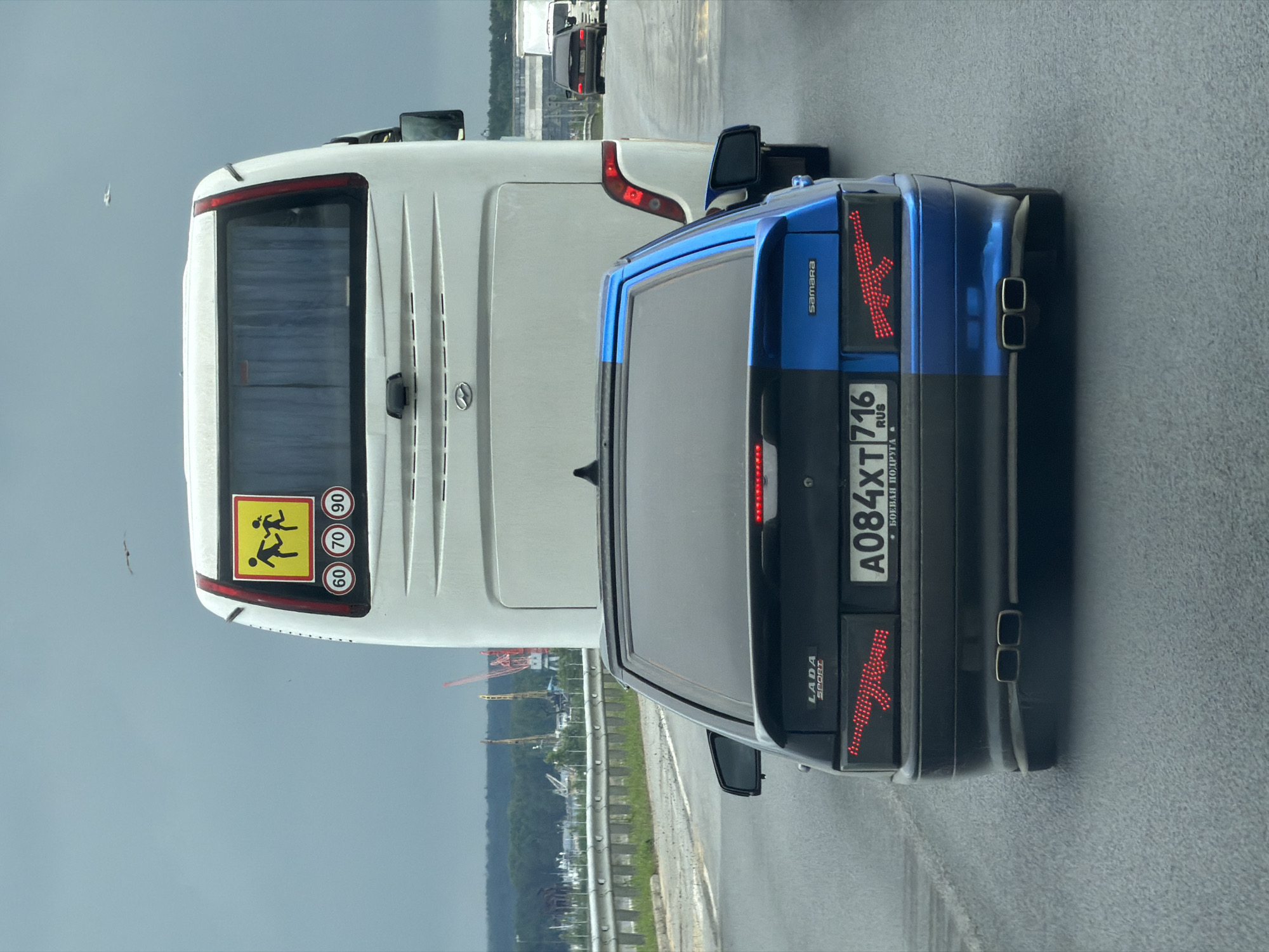 The image presents a rear view of vehicular traffic on a paved road under an overcast sky, in the city of Chaikovsky, Russia.
In the immediate foreground, a heavily modified Lada Samara sedan is visible. The vehicle's exterior is split vertically, with the left (driver's) side painted black and the right (passenger's) side painted blue. Emblems reading "LADA SPORT" and "Samara" are affixed to the trunk lid. The custom taillights illuminate in a pattern resembling an AK-47 assault rifle on both sides. The Russian license plate, A084XT 716 RUS, is centrally mounted, with the Cyrillic text "БОЕВАЯ ПОДРУГА" (Combat Girlfriend) positioned beneath it. The car features a dual exhaust system with two square tips on each side.
Directly ahead of the Lada, a white Hyundai passenger bus occupies the midground. The bus's rear window displays a yellow square sign depicting two running human figures, indicative of child transport, along with three circular speed limit signs marked "60," "70," and "90" km/h.
The background reveals a guardrail along the left side of the road, followed by a vegetated embankment. Beyond this, a wide body of water, likely a river, is visible. The far bank features an industrial landscape characterized by multiple large red and yellow cranes and various structures, interspersed with dense green trees. Further down the road, a white truck and a dark sedan are visible in the distance. The sky is uniformly grey, indicating overcast weather, and several small birds are observed in flight in the upper portion of the frame. No individuals are explicitly visible.