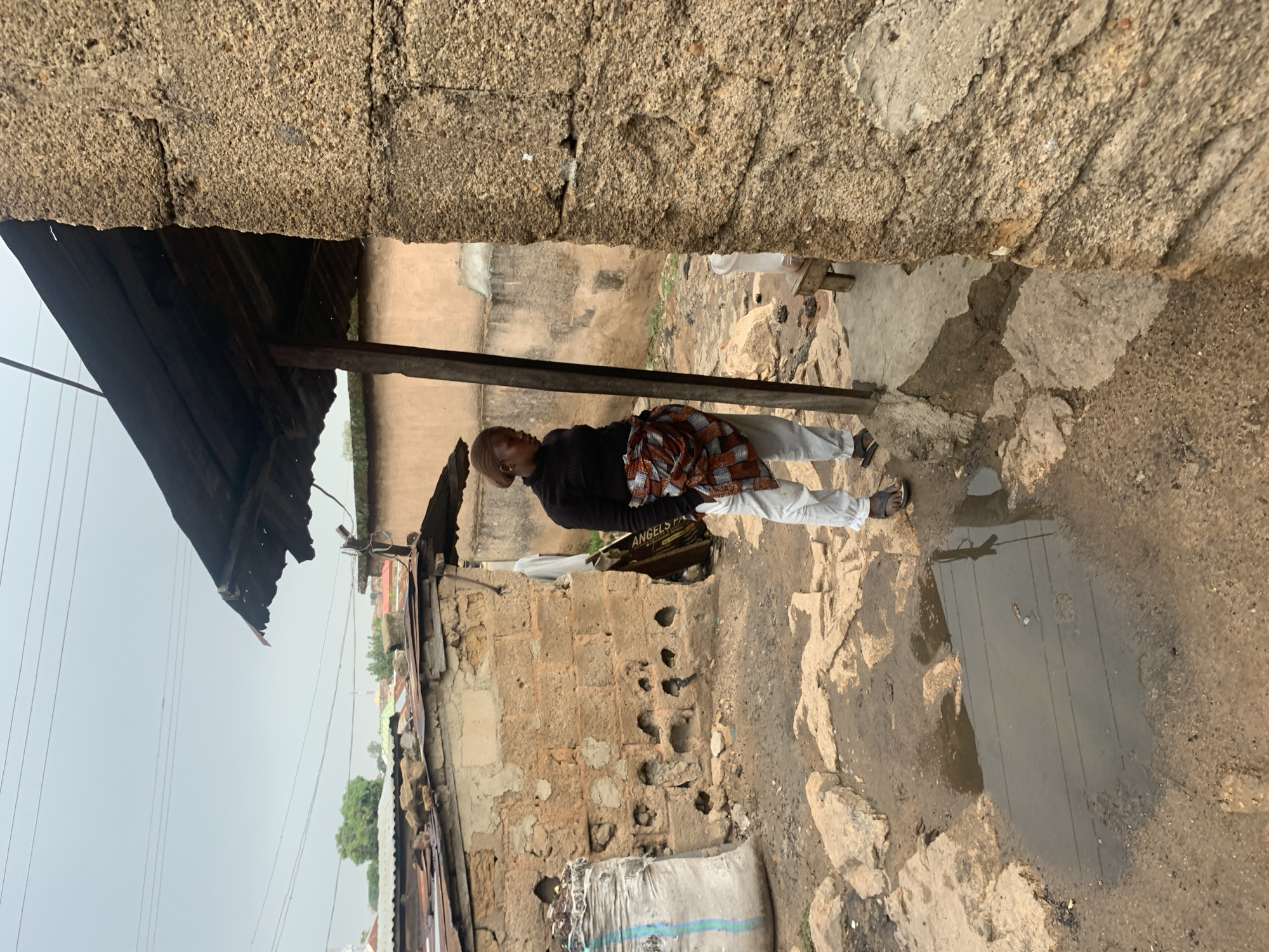 A vertical wide-angle shot depicts a narrow alleyway in Jos, Nigeria. The alley is framed by weathered, unfinished brick walls and a corrugated metal awning. The ground is a mix of dirt, stones, and a large puddle reflecting overhead power lines. A slender person wearing a dark long-sleeved shirt, light pants, and sandals stands in the alley. The person is carrying a patterned cloth over one shoulder. A partially visible bag is resting against one of the brick walls. Overhead, power lines stretch across a gray sky.