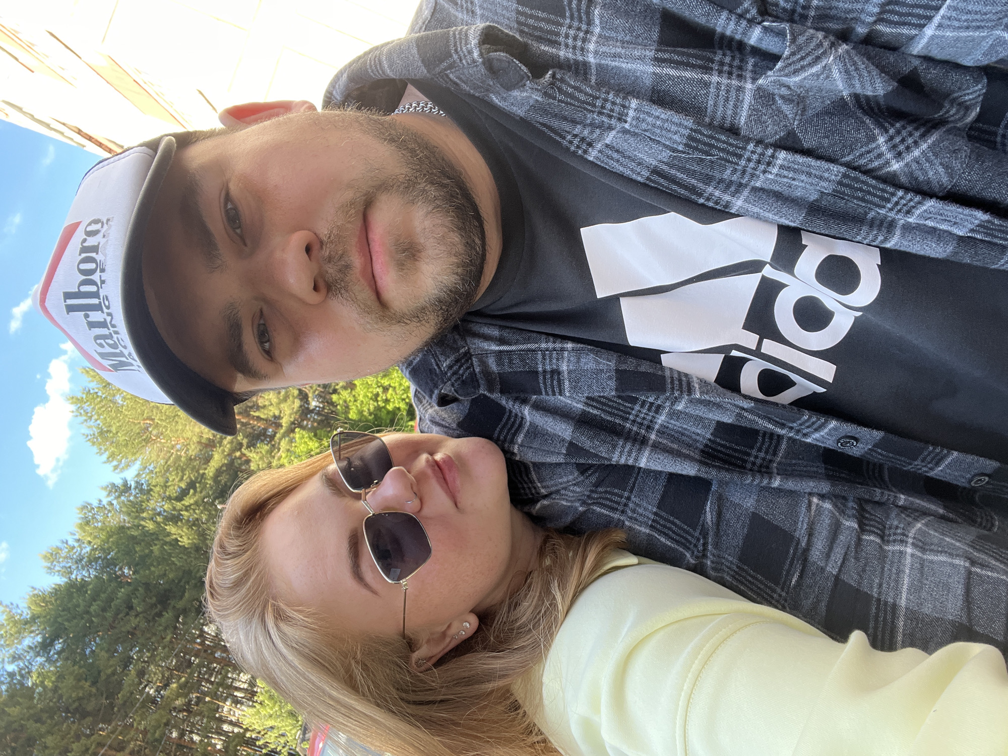 A selfie-style photograph captures a couple outdoors. The man, positioned at the top of the frame, wears a "Marlboro" branded cap, a black t-shirt with a white Adidas logo, and a grey plaid shirt. He has a beard and appears to be wearing a chain. The woman, situated below him, wears sunglasses and a yellow shirt. Trees and a blue sky are visible in the background. The location may be near trees. The location may be Chaikovsky, Russia, but there is no confirmation of this in the image. The couple appears to be posing for the photo.