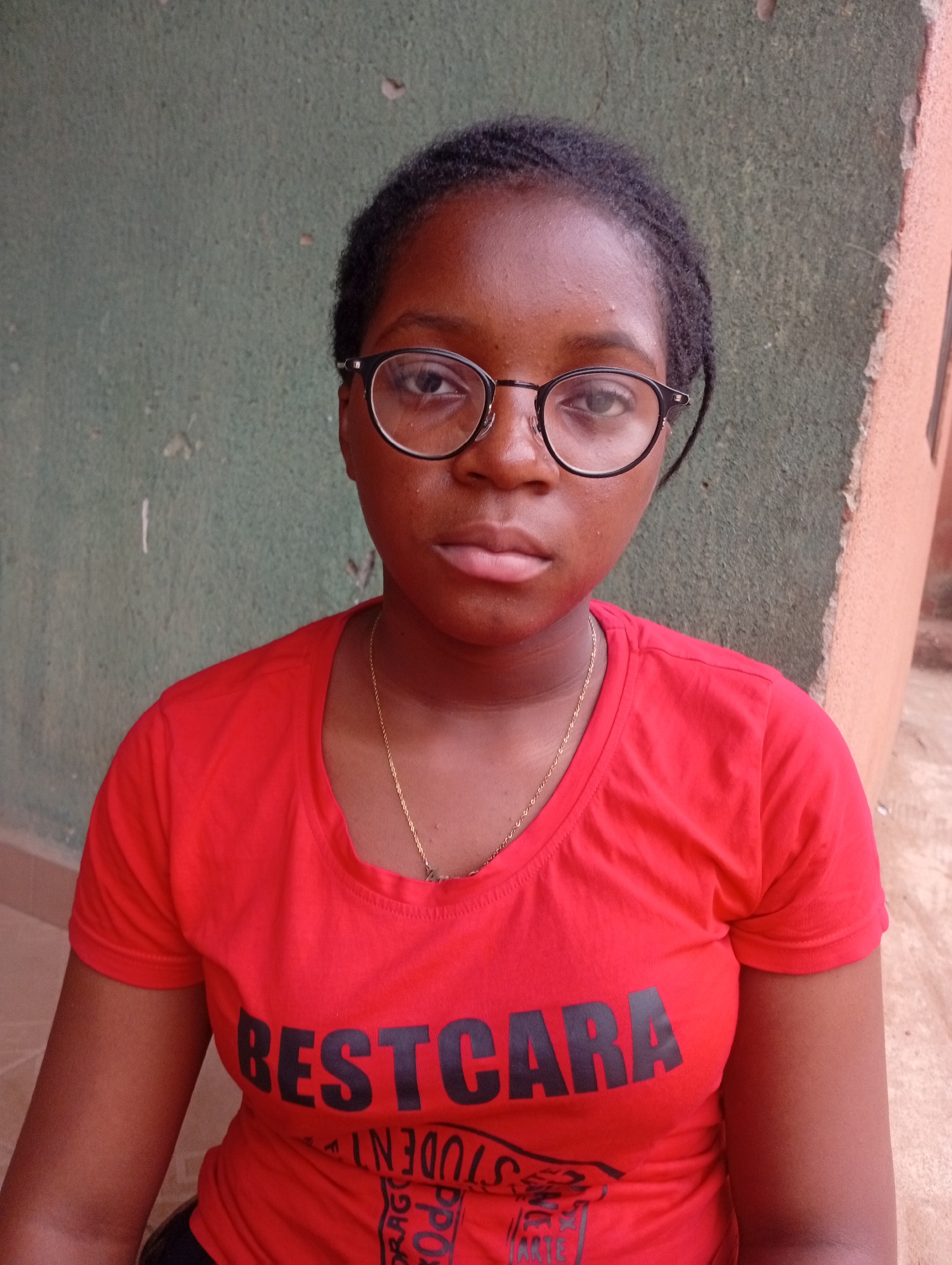 A young woman stands facing the camera, wearing black-rimmed glasses, a gold chain, and a red t-shirt with the word "BESTCARA" printed in black. She appears to be in Sabon Tasha, Nigeria, based on the background which includes a green wall with white marks and a section of orange wall. The woman's expression is neutral.