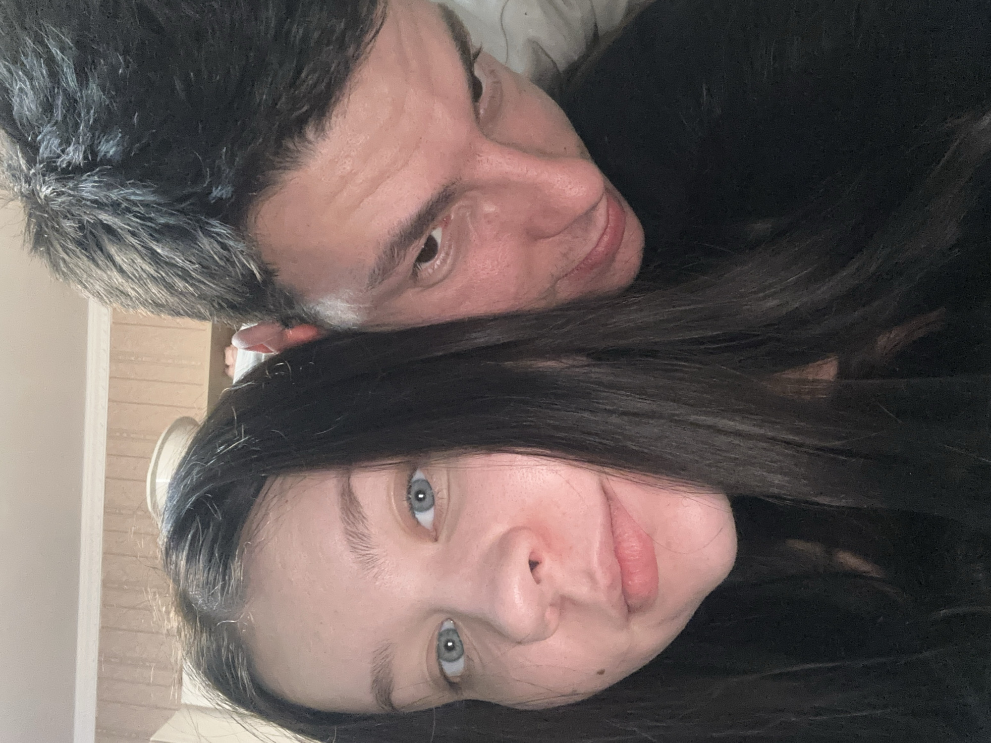 The image shows two people, a man and a woman, posing closely together for a selfie-style photograph. The man has short, dark hair with some graying and is positioned above the woman, looking downward with a slight smile. The woman has long, dark hair and is looking directly at the camera. The background is blurred and difficult to discern, but there is a hint of a wall and possibly a doorway in the upper left corner of the image. The location in Varna, Bulgaria cannot be determined from the provided image.