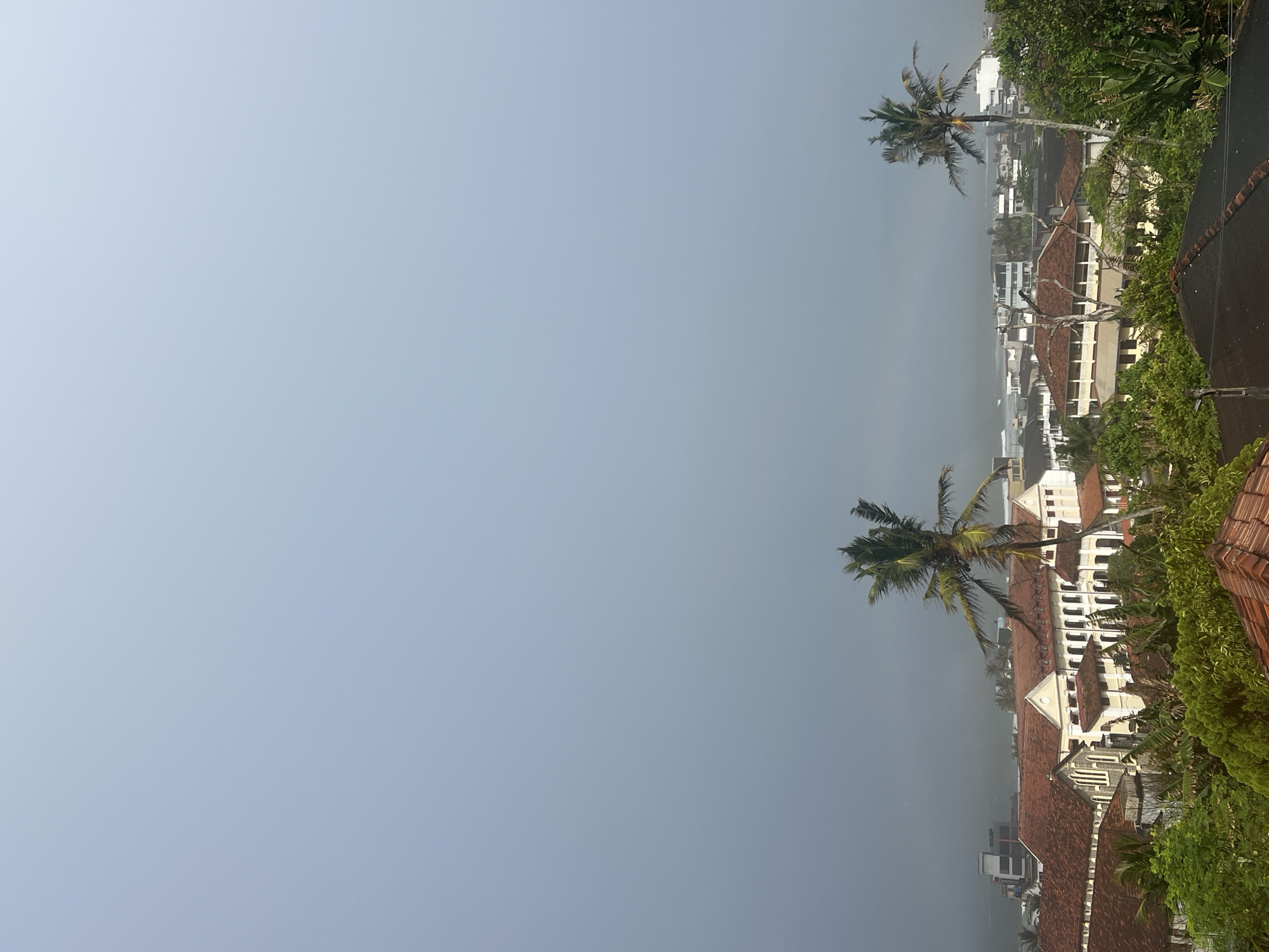 The image shows an aerial view of the city of Galle, Sri Lanka, on a hazy or overcast day. In the foreground, red-tiled roofs of buildings are visible, interspersed with lush green foliage. Two prominent palm trees stand tall among the vegetation. Behind the foreground, a collection of buildings with terracotta roofs and white or cream-colored walls are clustered together, suggesting a well-established urban area. Further in the distance, more modern buildings, including some with white facades, can be seen. The farthest part of the scene reveals the ocean, with waves breaking on a distant shoreline, indicating the city's coastal location. The sky is a uniform pale blue-gray, contributing to the overall muted atmosphere of the scene. There are no people visible in the image.