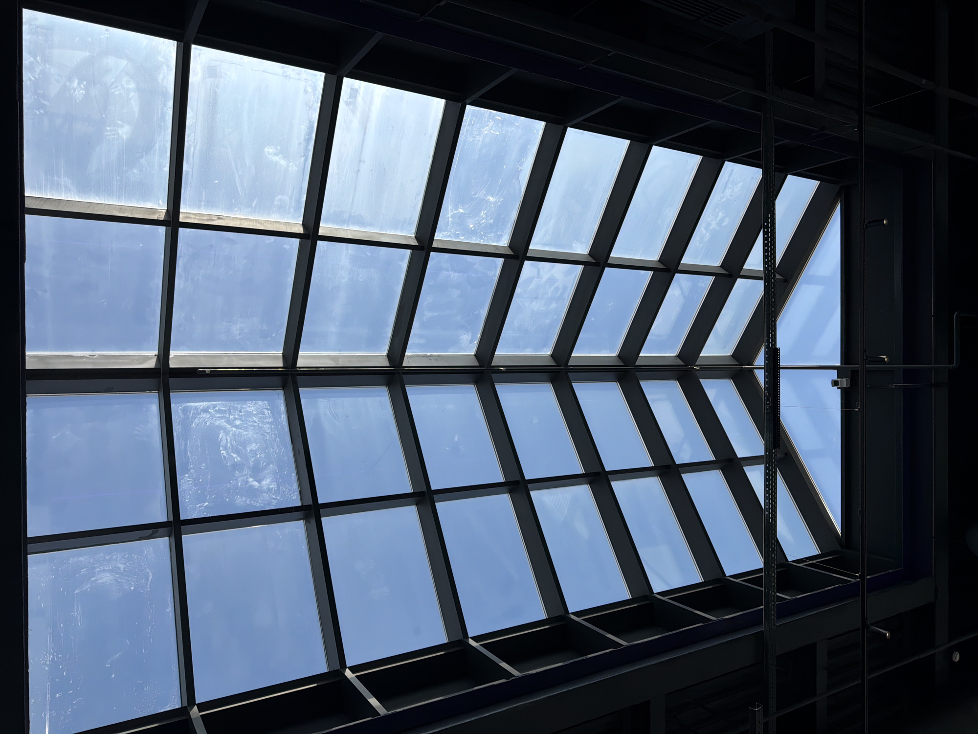 A low-angle shot reveals a multi-paneled glass ceiling, likely an atrium or skylight, filtering sunlight and offering a view of a blue sky with scattered clouds. The glass panels are framed by a dark, possibly black, grid-like structure. The right side of the frame includes additional dark metal structures and equipment, possibly ventilation or structural supports. The image was taken indoors, looking up. No people are visible. I am unable to confirm the location as Krasnoyarsk, Russia.