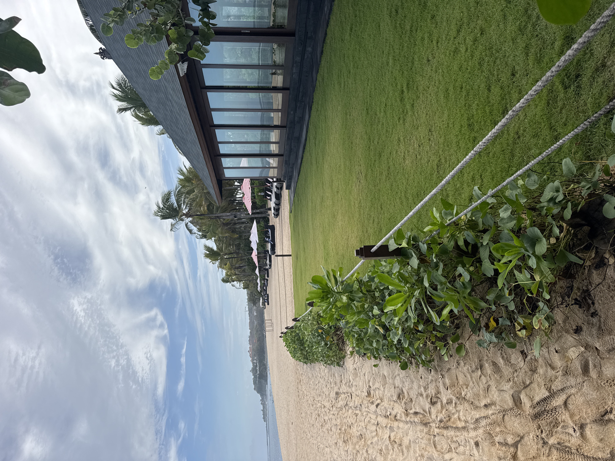 The image depicts a tranquil beach scene in Kuta Selatan, Indonesia, characterized by a clean sand beach, lush green grass, and a modern building with expansive glass windows. On the left, a sandy path shows footprints, suggesting recent activity, bordered by dense green foliage. A rope fence separates the sandy area from the manicured lawn on the right. In the distance, palm trees line the horizon, and beach umbrellas and lounge chairs are visible, indicating a resort or recreational area. The sky is partly cloudy with a blend of blue sky and white, wispy clouds. The overall impression is one of a serene tropical getaway.