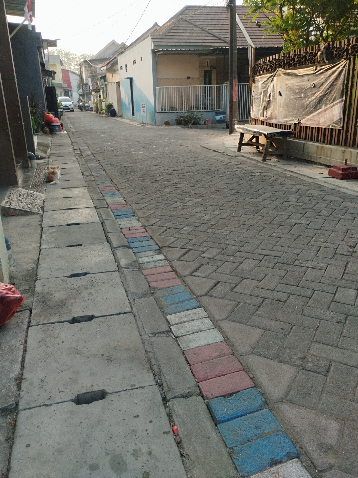 An outdoor, eye-level, medium shot captures a residential street in Surabaya, Indonesia, under an overcast sky. The foreground features a concrete drainage channel with visible openings, alongside a brick-patterned sidewalk with alternating red, white, and blue bricks. A ginger and white cat sits on the drainage cover facing left. The street itself is paved with gray interlocking bricks. In the mid-ground, a few houses with tiled roofs line the street, one with a white fence and plants. A motorcycle and a white car are parked further down the street. On the right side of the image, a wooden bench with a blue seat sits in front of a fence covered with a large, tattered tarp. The scene conveys a sense of quiet everyday life in a neighborhood.