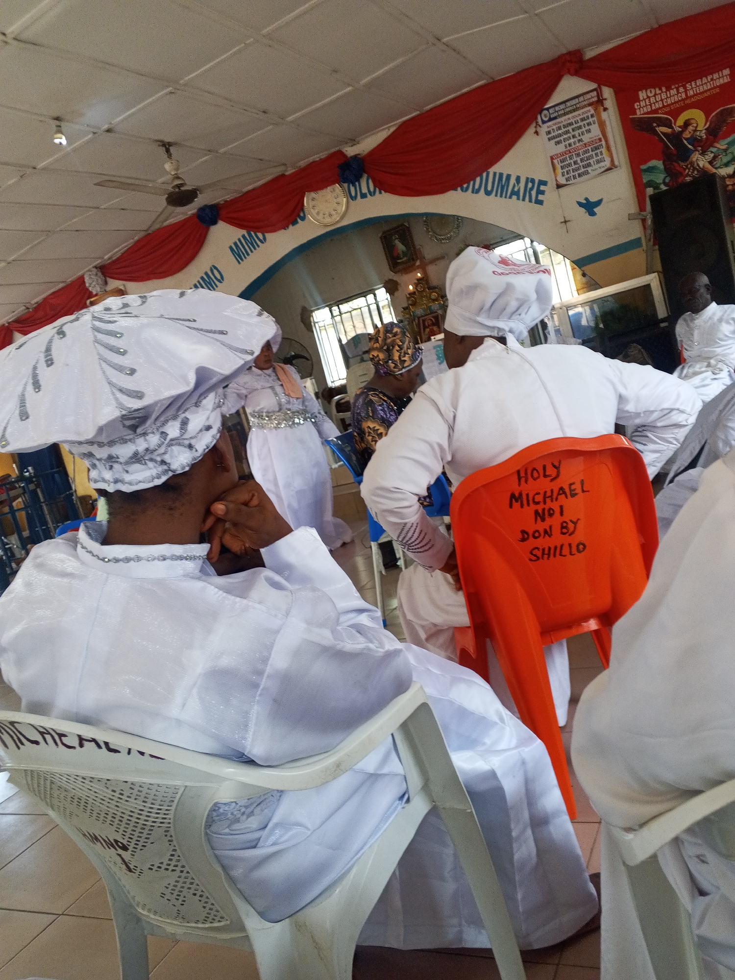 This image depicts an indoor religious gathering in the city of Lokoja, Nigeria. The scene is set within a hall or church sanctuary, characterized by light-colored walls, a tiled floor, and a suspended grid ceiling from which a ceiling fan and a single light bulb hang.
Multiple individuals, predominantly dressed in white religious attire, are present. In the immediate foreground to the left, a person is seated on a white plastic chair, viewed from behind. They wear a long-sleeved white garment adorned with silver chain-like embellishments on the collar and cuffs, paired with a large, elaborate white head covering featuring silver sequin or bead detailing. Their right hand is raised to their face, possibly resting their chin or covering their mouth.
To the right, slightly further back, another individual is seated on a bright orange plastic chair, also viewed from behind. They wear a plain white long-sleeved garment and a simple white cap. The backrest of the orange chair bears the handwritten inscription in black marker: "HOLY MICHAEL Nº 1 DON BY SHILLO."
In the mid-ground, towards the left, another person in white attire with a silver beaded belt and a pale pink or peach sash stands facing away from the camera. Further into the background, several other people are visible, mostly dressed in white. One person on the far left has a patterned headwrap. On the far right, a person in white is partially visible.
The walls are decorated with red fabric drapes secured by blue rosettes. A light blue curved section of the wall features white text, "MIMO MIMO LOLO ODUMARE" (likely Yoruba for "Holy Holy Lord Odumare"). A round wall clock is positioned on the draped wall. To the right, banners are displayed: one features a depiction of an angel with a sword and wings, along with text including "HOLY CHERUBIM & SERAPHIM BRDG AND CHURCH INTERNATIONAL" and "OGUN STATE HEADQUARTER." Another smaller sign displays a blue dove symbol above a cross. A large black speaker box is situated on the far right.
In the background to the left, an altar or shrine-like arrangement is visible, featuring religious icons, candles, and a golden candelabrum. Windows with security bars allow natural light into the space. The overall environment suggests an active religious service or event.