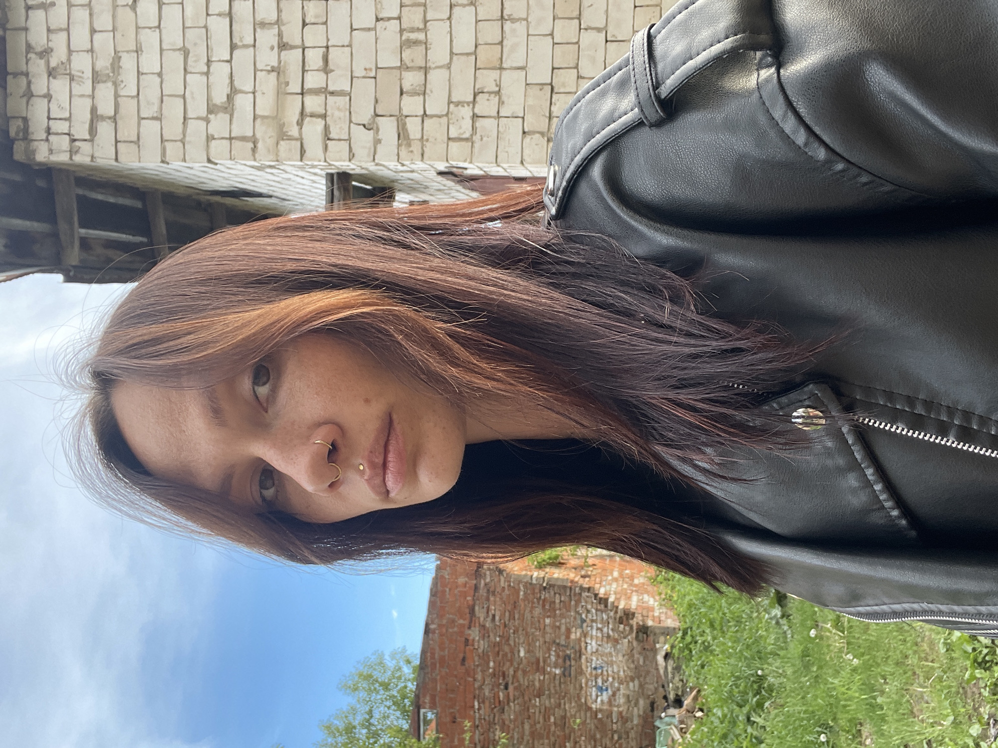 A selfie of a person with auburn hair and fair skin, likely female, is positioned against a backdrop of dilapidated brick buildings and greenery. The person is wearing a black leather jacket and has piercings on their nose and lip. The background features a brick building with multiple white brick blocks, and another building with red bricks; grass, and a tree. The sky is blue and partially visible. The location is suggestive of a decaying urban area, possibly Oktyabr'skii, Russia.