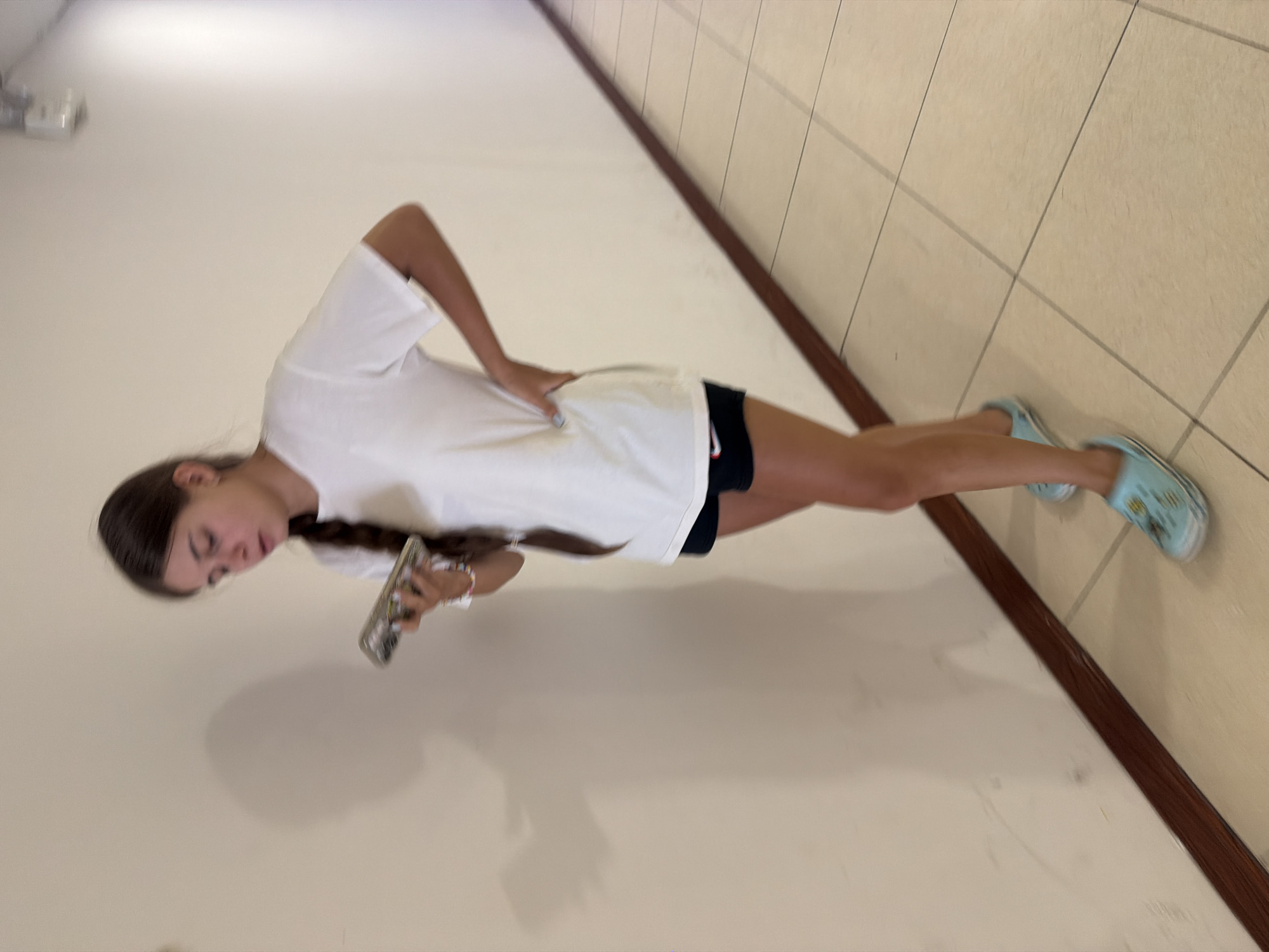 A young woman with a braid stands in a hallway, looking at her phone. She is wearing a white t-shirt and black shorts. She has light blue shoes on her feet. The hallway has a tiled floor and a wall with a shadow cast on it. The location is identified as a hallway in Nha Trang, Vietnam.
