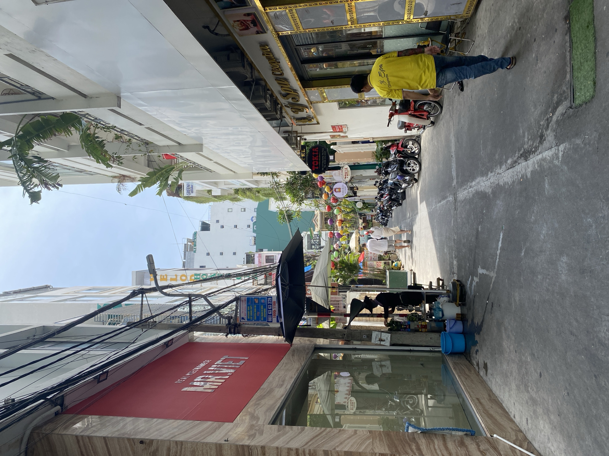 This is a sideways photograph of a street scene in Nha Trang Ward, Vietnam. The image captures a bustling urban environment with buildings lining a narrow street. Several motorbikes are parked along the side of the road, suggesting it is a common mode of transportation.
In the foreground, the side of a building with a red awning is visible, displaying the text "REAL VIETNAMESE". Above this, a network of electrical wires creates a dense, tangled overhead canopy.
Further down the street, there are storefronts with signs indicating various businesses, including "TOKIA" and "Spa Cleopatra". Colorful lanterns are strung across the street, adding a festive touch. People are walking along the sidewalk, and one person wearing a bright yellow shirt is interacting with a parked red motorbike. Another individual in white clothing is also visible on the sidewalk.
The scene appears to be taking place during daylight hours, with bright sunlight casting shadows. The overall atmosphere is lively and typical of a commercial street in a Vietnamese city. The presence of multiple businesses, parked vehicles, and pedestrians indicates an active urban area.