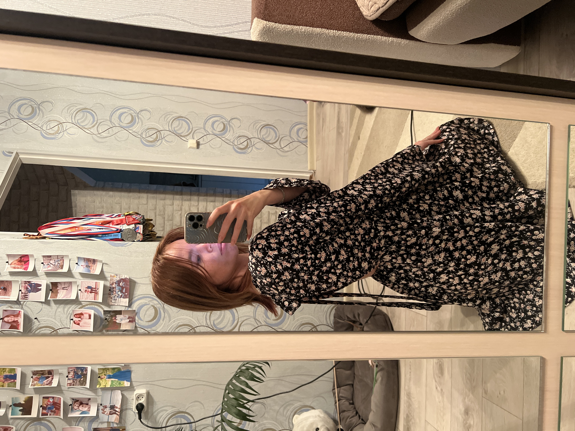 A young woman with auburn hair is kneeling in front of a mirror, taking a selfie with her phone. She is wearing a black dress with a floral print. The mirror reflects a room with patterned wallpaper, framed photographs on the wall, and a collection of medals hanging near the top of the frame. To the left, a plant is visible in a pot, and a pet bed is on the floor. The floor appears to be light-colored wood or laminate. The room is likely a residential setting, possibly a bedroom or living area, in Chaikovsky, Russia.