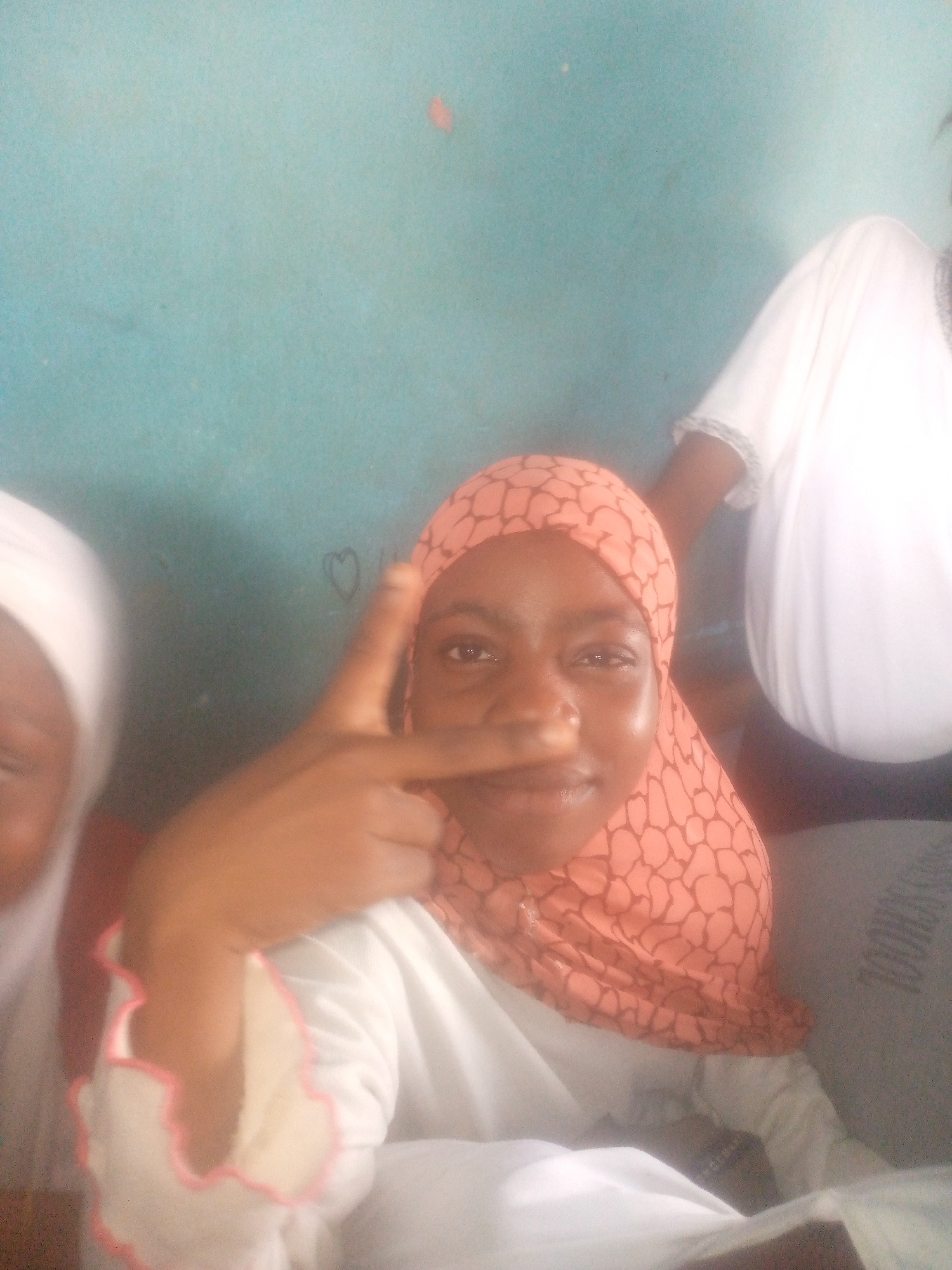 Here is a forensic description of the image:
**Scene:** Indoor setting, possibly a classroom, given the light blue wall and the presence of multiple individuals. Location: Unknown, Unknown.
**People:**
* **Subject 1:** A female wearing a peach-colored patterned hijab and a white garment with pink edging on the sleeves. She is making a "V" sign with her fingers and holding it near her nose.
* **Subject 2:** Partial view of a person to the left, wearing a white head covering.
* **Subject 3:** Partial view of a person to the right, wearing a white garment.
* Possible additional individuals are implied but not clearly visible.
**Objects:**
* Light blue wall with a small heart drawn on it.
* White garments worn by at least three people.
* Hijab worn by Subject 1.
* Possibly a table or desk area in front of the visible individuals.
**Actions:**
* Subject 1 is posing and making a "V" sign.
* Other individuals appear to be present but their actions are not clear from the image.
**Interactions:** No clear interactions between individuals are evident, but the group setting suggests a shared activity or environment.
**Environment:** The room is lit with natural or artificial light, given the brightness and lack of harsh shadows. The condition of the wall suggests a well-used or slightly aged space.