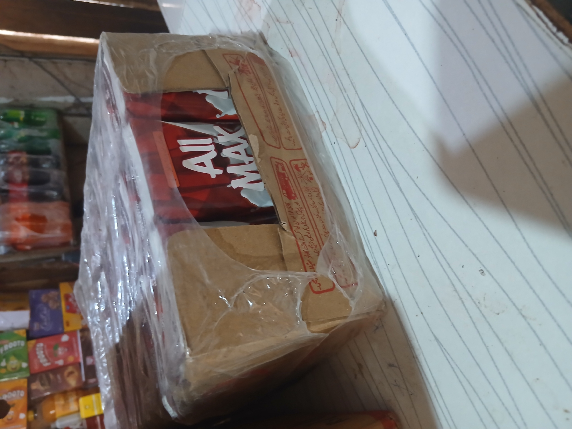 The image shows a close-up, slightly angled view of a cardboard box wrapped in clear plastic. The box contains multiple red cartons with the text "All MAX" visible in white lettering on a red background. Some of the cartons are partially obscured by the plastic wrap and the box itself. The box is sitting on a white surface with gray striped lines, possibly a table or counter. In the background, on a shelf, are various products, including bottles of soda and other packaged goods. The setting appears to be a shop or storage area, likely within Peshawar City Tehsil, Pakistan, based on the context of the request. There are no people visible in the image. The lighting is moderate, casting some shadows. The overall condition of the items appears to be typical for retail or storage.