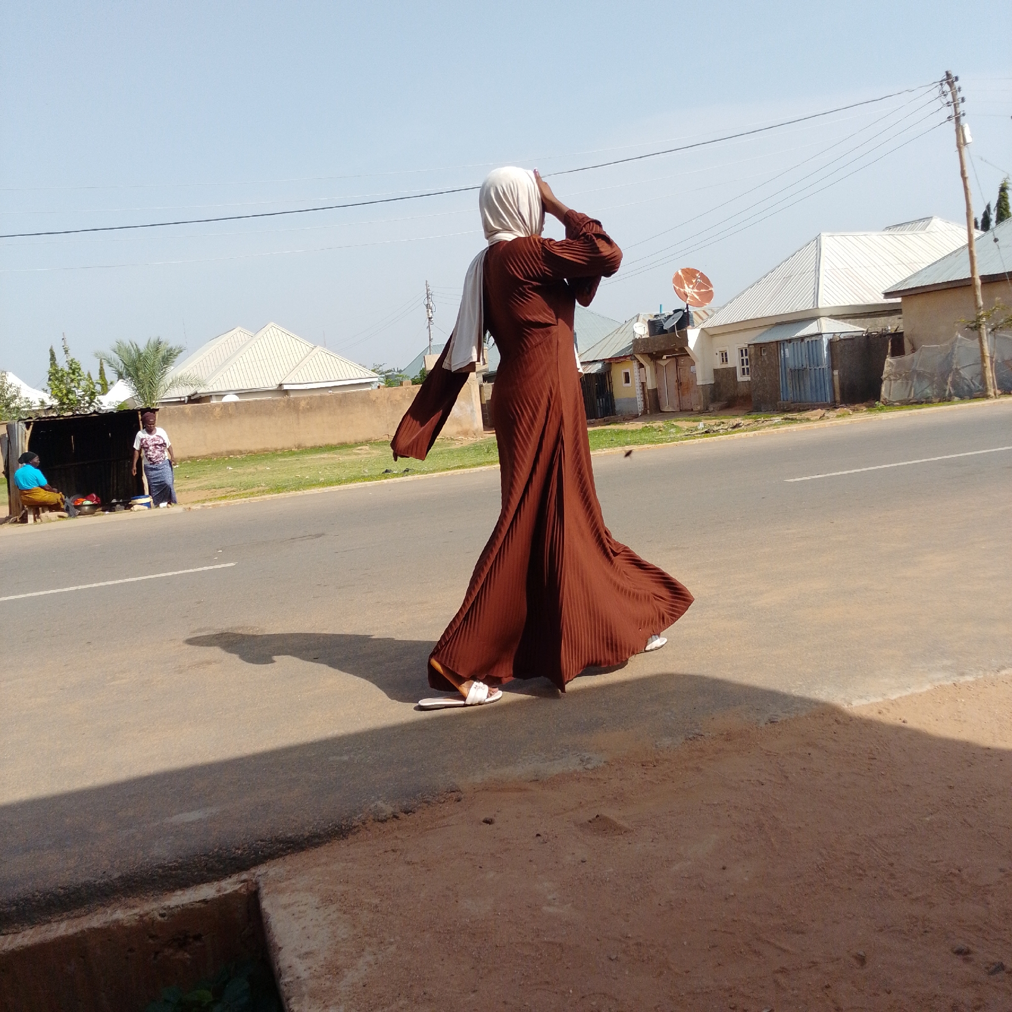 In Gombe, Nigeria, a woman wearing a flowing brown abaya and a white hijab stands on a paved road. Her hand is raised to her forehead, seemingly shielding her eyes from the sun. The abaya has a pleated design, and she is wearing white sandals. Behind her, a residential area is visible with several buildings featuring white, corrugated metal roofs. A red satellite dish is mounted on one of the buildings. In the distance, two other individuals are seen near a roadside stall. One person, wearing a blue top and yellow skirt, is seated on a stool, while another individual in a white t-shirt and patterned skirt stands near a display of items. The environment appears to be a sunny day with a clear sky.