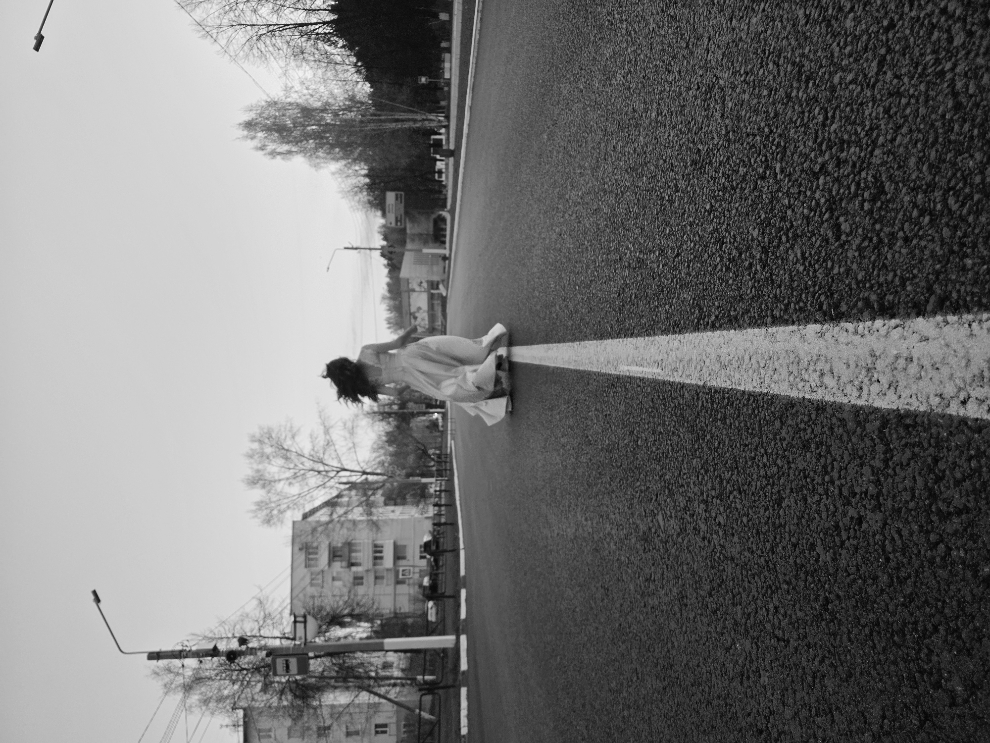 A grayscale, low-angle shot shows a person in a light-colored dress leaning over on a road in Chaikovsky, Russia. The road is asphalt with a painted white line. Buildings and trees line the side of the road. A lamppost is visible in the lower left of the frame. The sky is overcast.