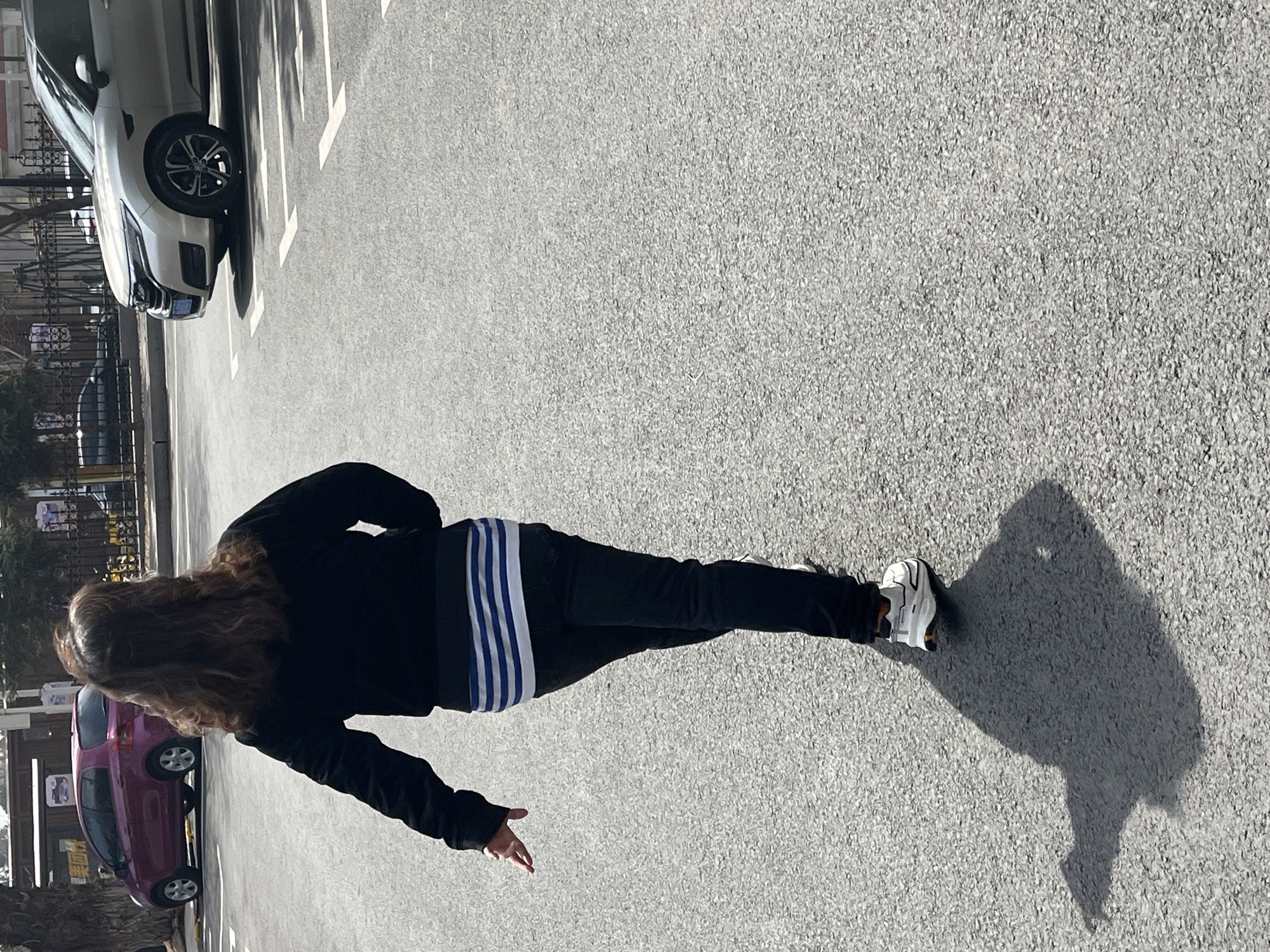 A woman with long curly brown hair, wearing a black jacket and black pants with blue stripes, is walking on a paved surface. Her right hand is outstretched. She is wearing white sneakers. Her shadow is visible. Behind her is a parking lot with several cars, including a white car and a purple car. Trees are visible in the background. The scene is located in Changchun City, China.