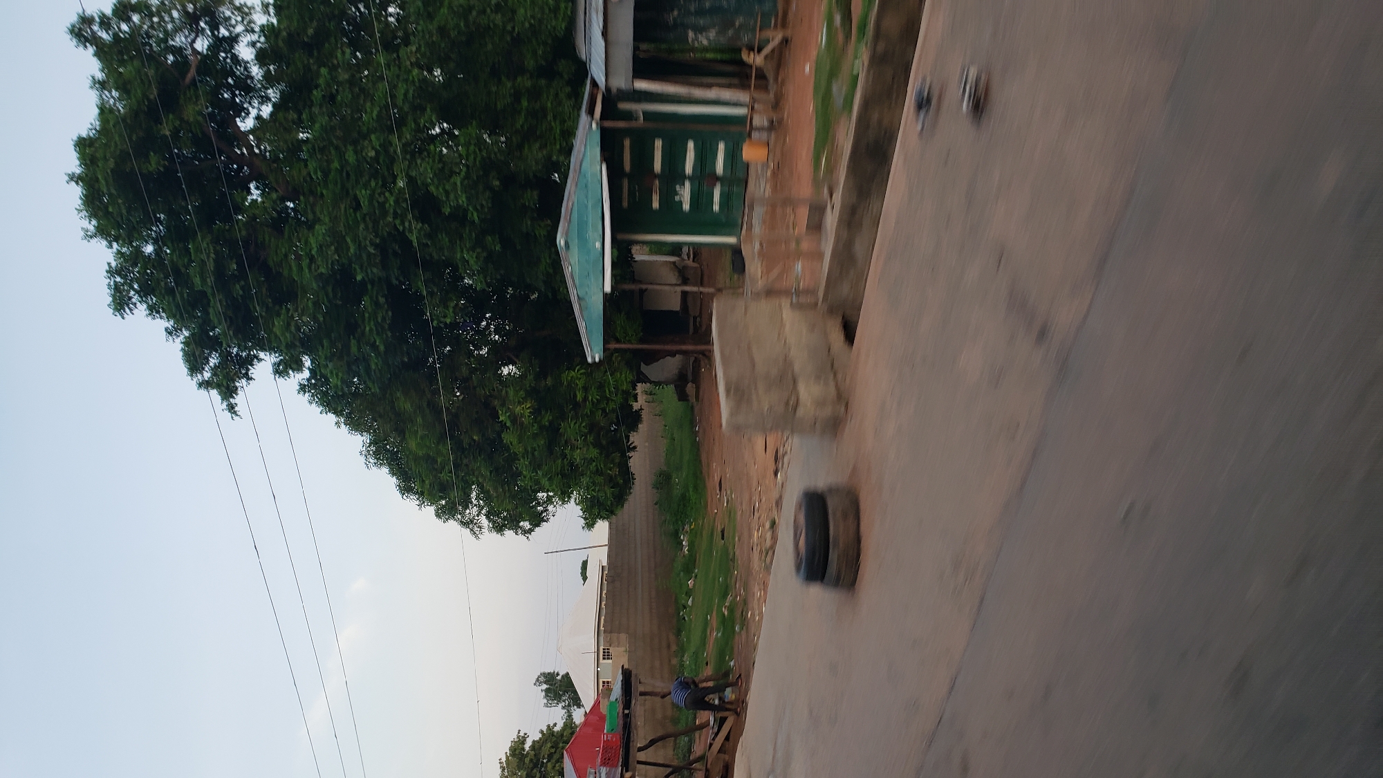 A low-angle, slightly blurred medium shot shows a street scene in Jalingo, Nigeria, under a pale sky. In the foreground, a paved road is visible with a drainage channel running along its right edge. Two stacked tires lie on the road to the left of the frame. In the middle ground, a man in a striped shirt and dark pants is bent over, appearing to be working with a rake on a dirt area. To the right of the man is a small, green structure with a light-colored roof, resembling a shipping container that has been converted into a stall, with an overhang supported by posts. Behind this structure is a large, leafy tree. Further back, a brick wall separates the immediate area from a residential building with a white, gabled roof. Overhead, three power lines stretch across the sky. The scene suggests a roadside market or informal settlement.