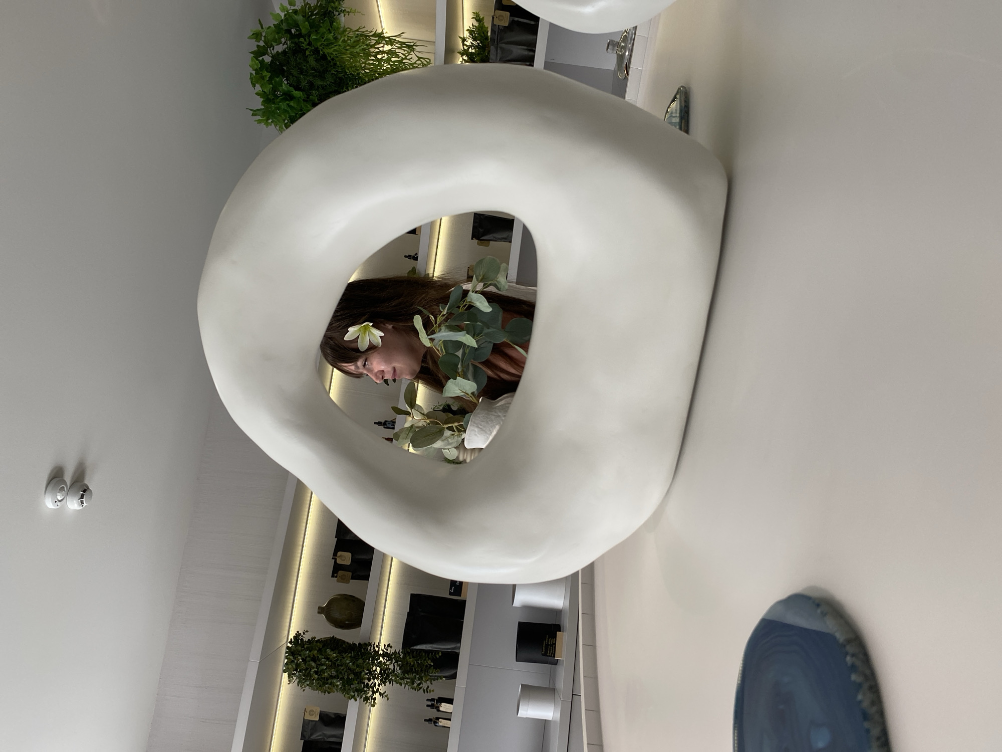 The image depicts an indoor scene, likely a retail space, with a minimalist aesthetic. In the foreground, a large, abstract, white sculpture with a hole in the center dominates the view. A woman with long brown hair and a flower tucked behind her ear is visible through the sculpture's opening. She is holding a small potted plant with green leaves. The background shows shelves stocked with various items, including black bags, bottles, and decorative vases, illuminated by soft strip lighting. There is also a faux potted plant with lush green foliage on a shelf. In the upper left corner of the image, a white smoke detector is attached to the ceiling. The overall environment appears clean and well-lit, suggesting a modern and curated space. The image does not contain any information that could place it in Votkinsk, Russia.