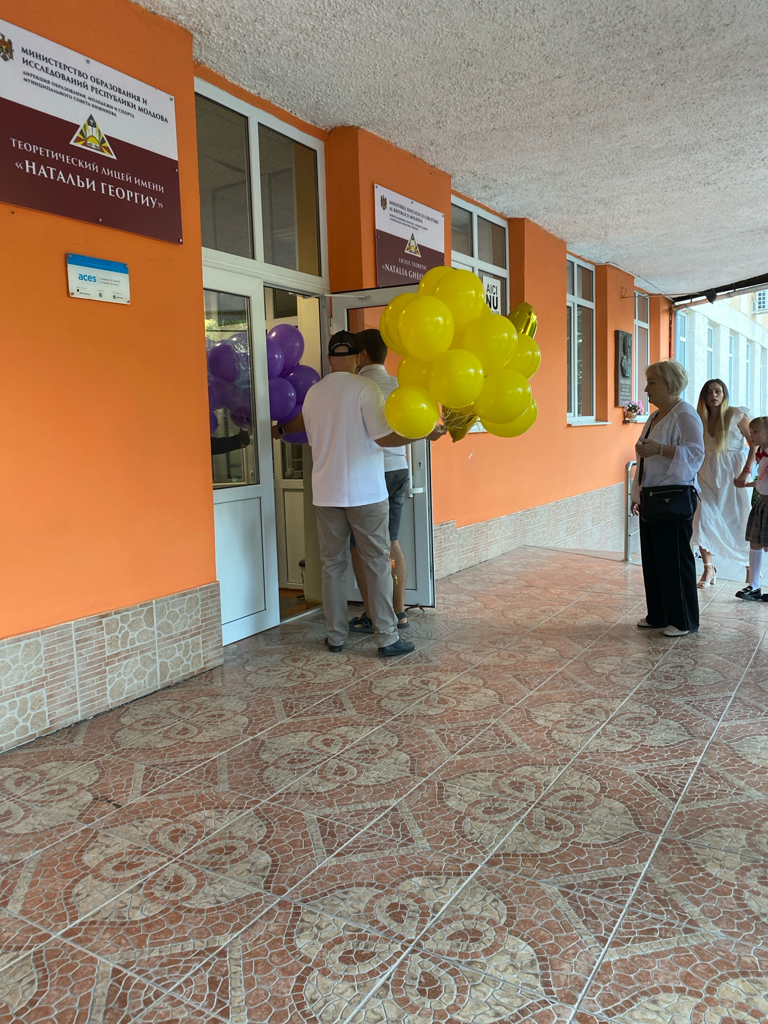 The image depicts the entrance to the Theoretical Lyceum named "Nataliya Gheorghiu" located in the city of Chișinău, Moldova. The building's exterior features an orange facade, with tiled flooring in the walkway. Several people are present, including men carrying bunches of yellow and purple balloons, suggesting a celebratory event such as the start of the school year. A woman in a white top and black pants stands to the right, observing the scene, while other individuals, including young girls, are partially visible in the background. Signage on the building indicates it is under the Ministry of Education and Research of the Republic of Moldova. The overall atmosphere appears to be one of anticipation and festivity.