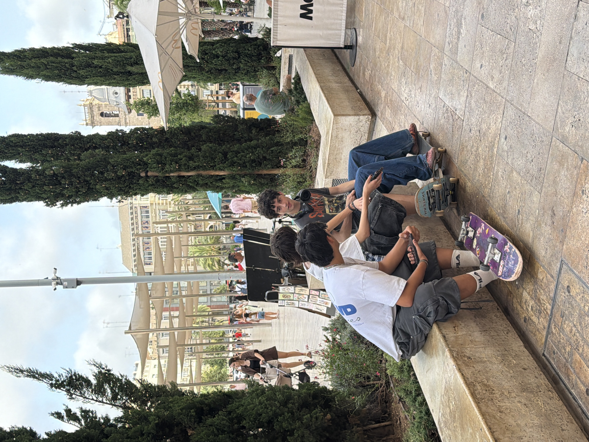 In the city of Unknown, Unknown, on a cloudy day, three young individuals are seated on a textured stone bench. The person on the left wears a white t-shirt and dark cargo shorts, with white socks that have a red and blue logo. A purple skateboard rests near their feet. The middle individual is dressed in a grey t-shirt and dark shorts, holding a smartphone. A blue skateboard is positioned in front of them. The person on the right wears a dark t-shirt, blue jeans, and red shoes, holding a phone and seated on a skateboard. Behind them, other people are visible in the background, walking through a plaza with cypress trees and architectural structures. One individual pushes a stroller, and a person in a green shirt is seated on a planter. White umbrellas and awnings are present, suggesting an outdoor public space with amenities.