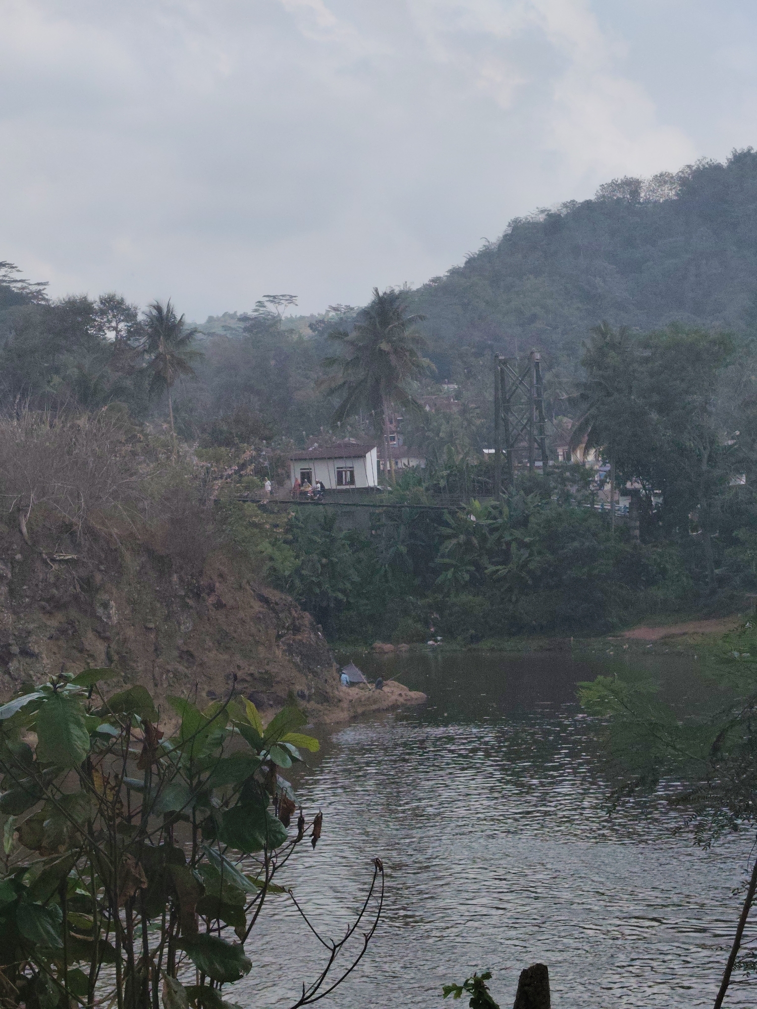 The image depicts a cloudy, overcast day scene with a body of water in the foreground. A rocky embankment rises steeply on the left, covered with dry vegetation. On the right side of the water, lush green trees and foliage create a dense backdrop.
In the mid-ground, a small bridge crosses the water, and on top of it, there are several buildings, including a white house with a red roof and dark-trimmed windows. A few people are visible near the buildings and on the bridge. Further back, a metal pylon structure stands amongst the trees, suggesting infrastructure like a power line or another bridge. The landscape transitions into a forested hill that dominates the background, disappearing into the hazy sky.
A single person is seated on a rock near the water's edge on the left side. The water's surface is rippled, indicating a light breeze. In the immediate foreground, branches and leaves of green plants are visible.
This scene is identified as located in Tasikmalaya, Indonesia.
