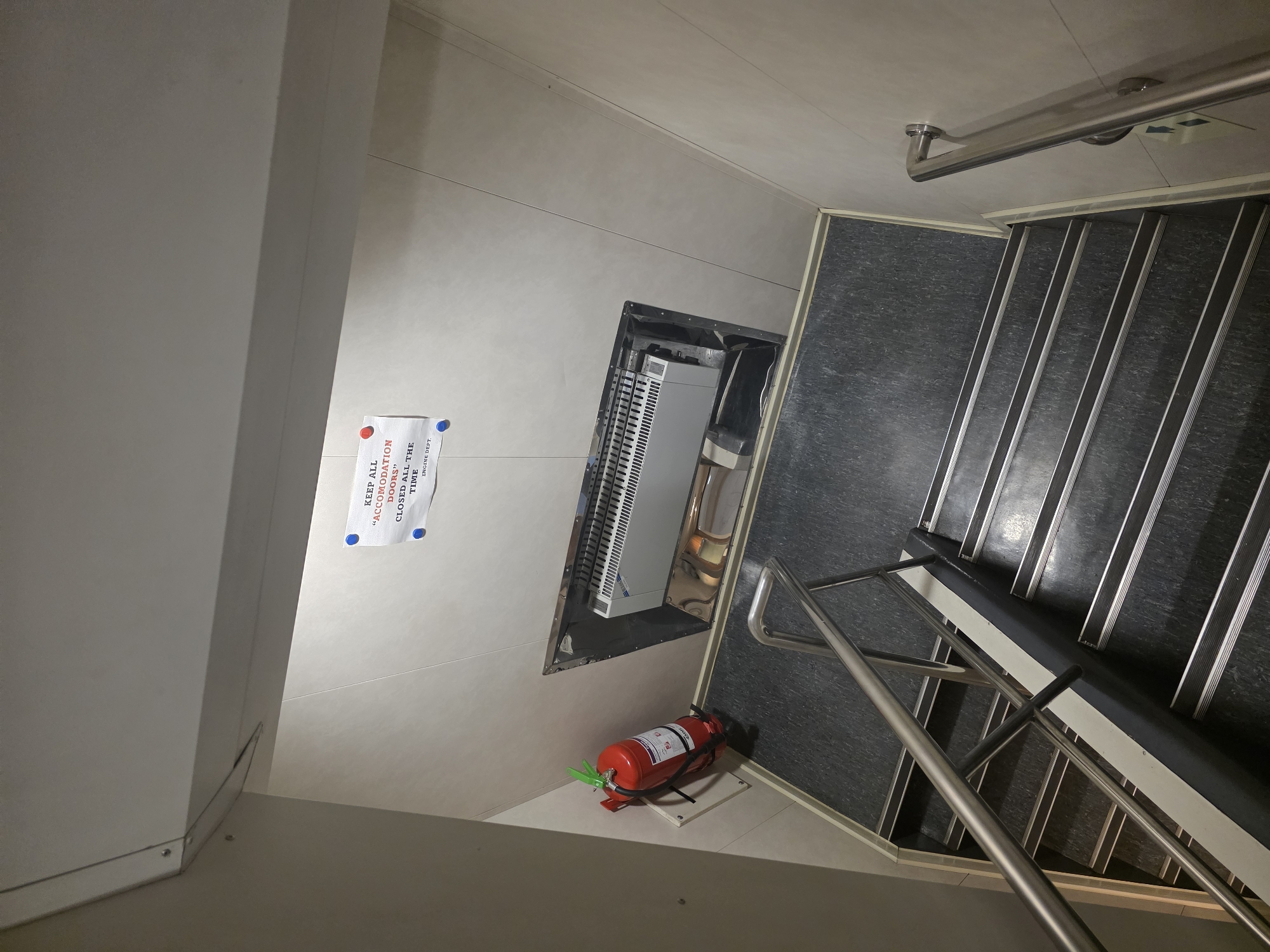 The image shows a top-down view of a dimly lit staircase with dark grey flooring and metal railings. On the wall to the left, there is a red fire extinguisher. Above the stairs, a sign is attached to the wall with two red pushpins and one blue pushpin. The sign reads: "KEEP ALL "ACCOMODATION DOORS" CLOSED ALL THE TIME ENG. DEPT." In the wall above the stairs, there is a recessed area containing a white and silver HVAC unit. The walls are light-colored and appear to be paneled. The overall scene suggests an interior, possibly on a ship or in a utilitarian building, given the presence of a fire extinguisher and a technical sign. There is no indication of the city of Brindisi, Italy, in the image.