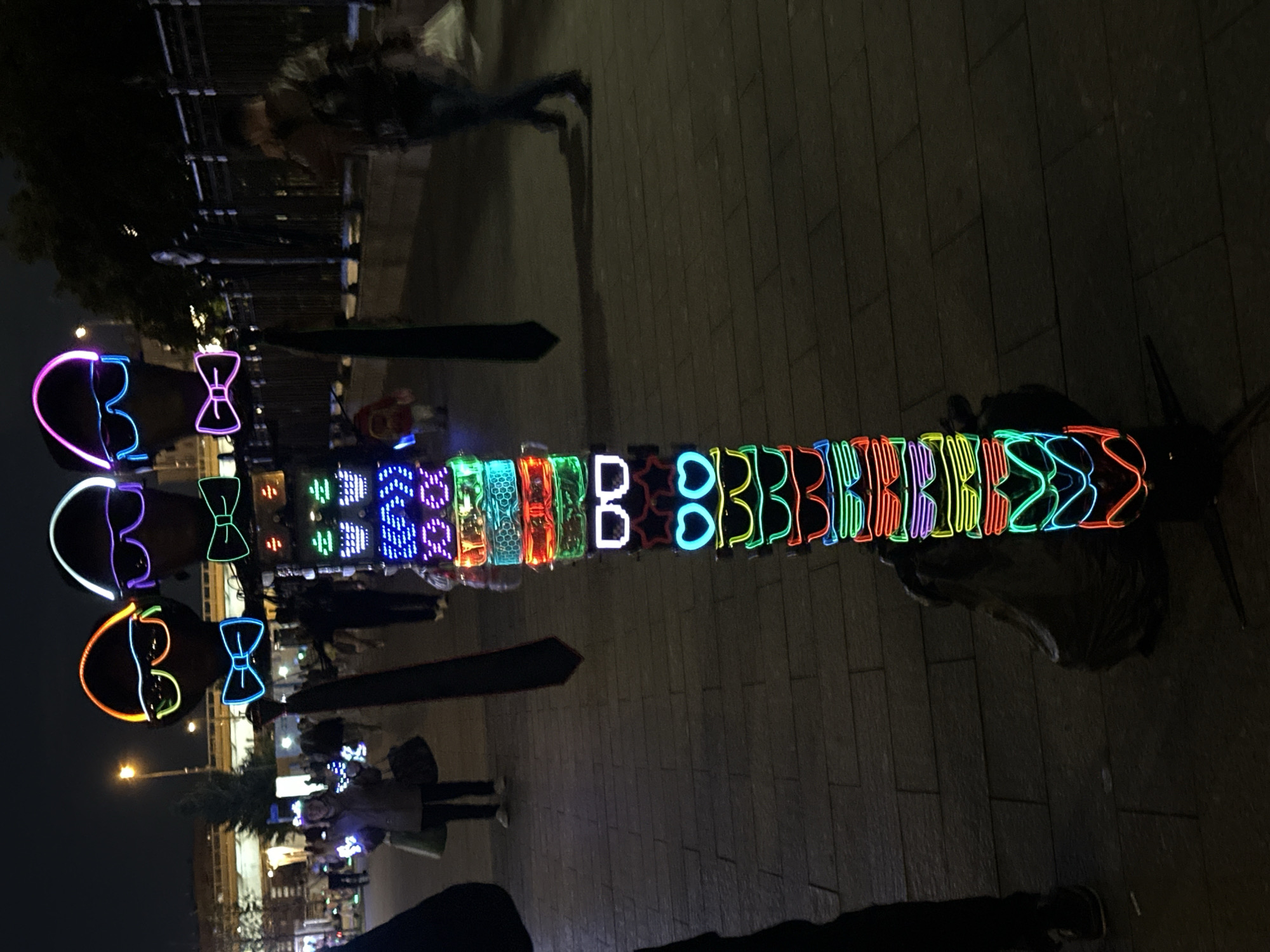 The image depicts an outdoor night scene in Moscow, Russia, capturing a vibrant display of glowing novelty items. The perspective is from a low angle, looking towards a colorful, elongated stand of illuminated objects.
The central focus is a long, horizontal arrangement of various neon-style lights. On the left side of this display, three sets of glowing "sunglasses" or masks are prominently featured, each paired with a glowing "bow tie." These are in shades of purple, orange, and green, with corresponding purple, blue, and green bow ties. To the right of these, the display continues with a sequence of individual glowing shapes and letters. Visible patterns include illuminated squares, hexagons, stars, and heart shapes, interspersed with repeated letter-like forms, most clearly showing multiple 'B's. These elements glow in a rainbow of colors including blue, green, red, orange, yellow, and purple.
The scene is set in a public area, possibly a street or market, with paved ground visible beneath the display. In the background, other people can be seen walking or standing, suggesting a lively evening atmosphere. One person in a light-colored jacket stands near the display, while another blurred figure walks past further back. Dim streetlights and the outlines of distant buildings and trees are visible against the dark night sky. The overall impression is one of entertainment and festivity, with the bright lights contrasting sharply with the dark surroundings.