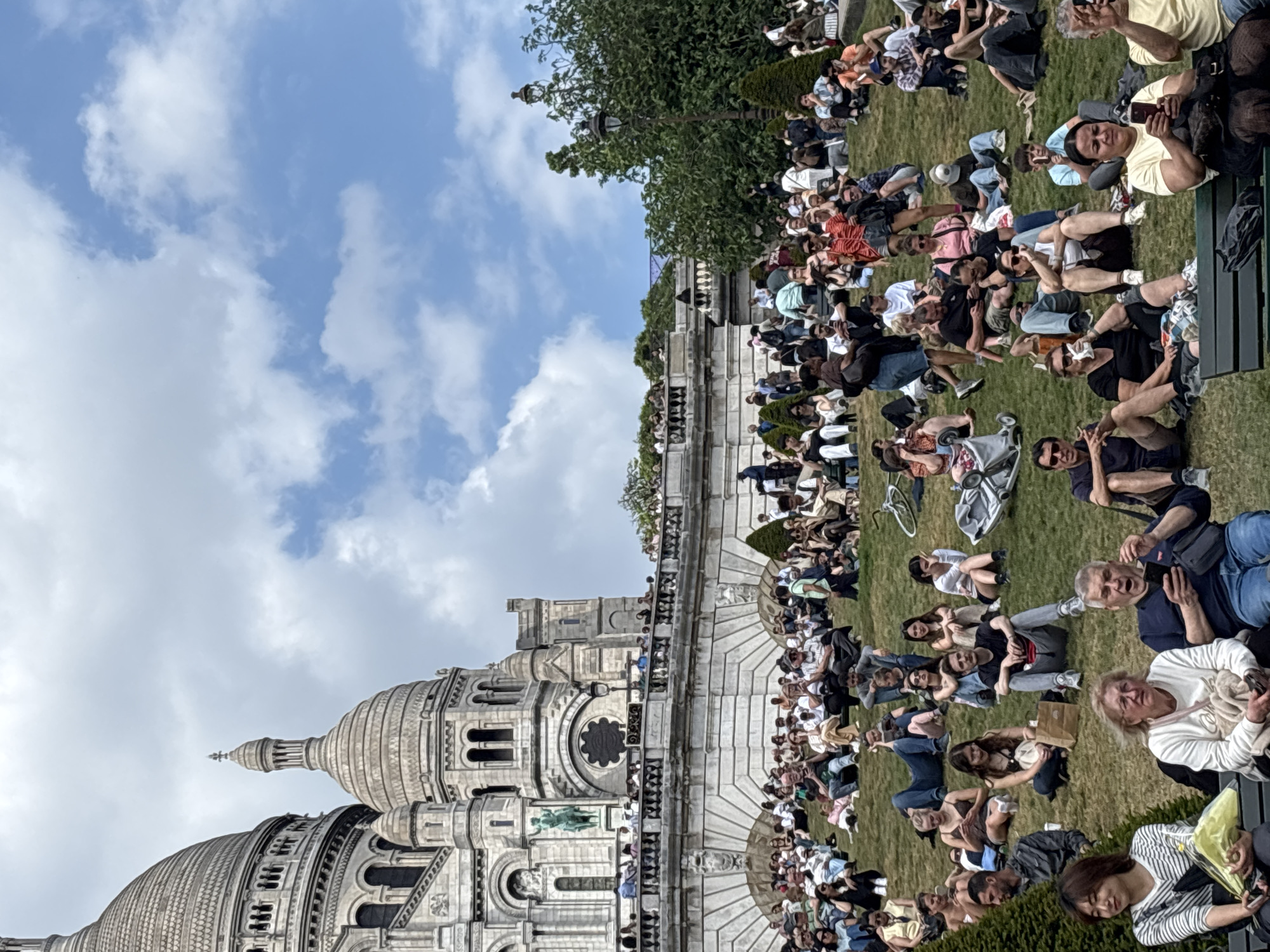 The image depicts a large group of people gathered on the lawn in front of the Sacré-Cœur Basilica in Paris, France. The basilica, a prominent landmark, occupies the left side of the frame, showcasing its architectural details. The people are mainly sitting or lounging on the grass, appearing relaxed and enjoying the environment. Some are using their phones, while others are conversing or simply observing the surroundings. The sky is partly cloudy, suggesting a pleasant day. The environment is filled with tourists and possibly locals enjoying an outdoor experience in the heart of Paris.