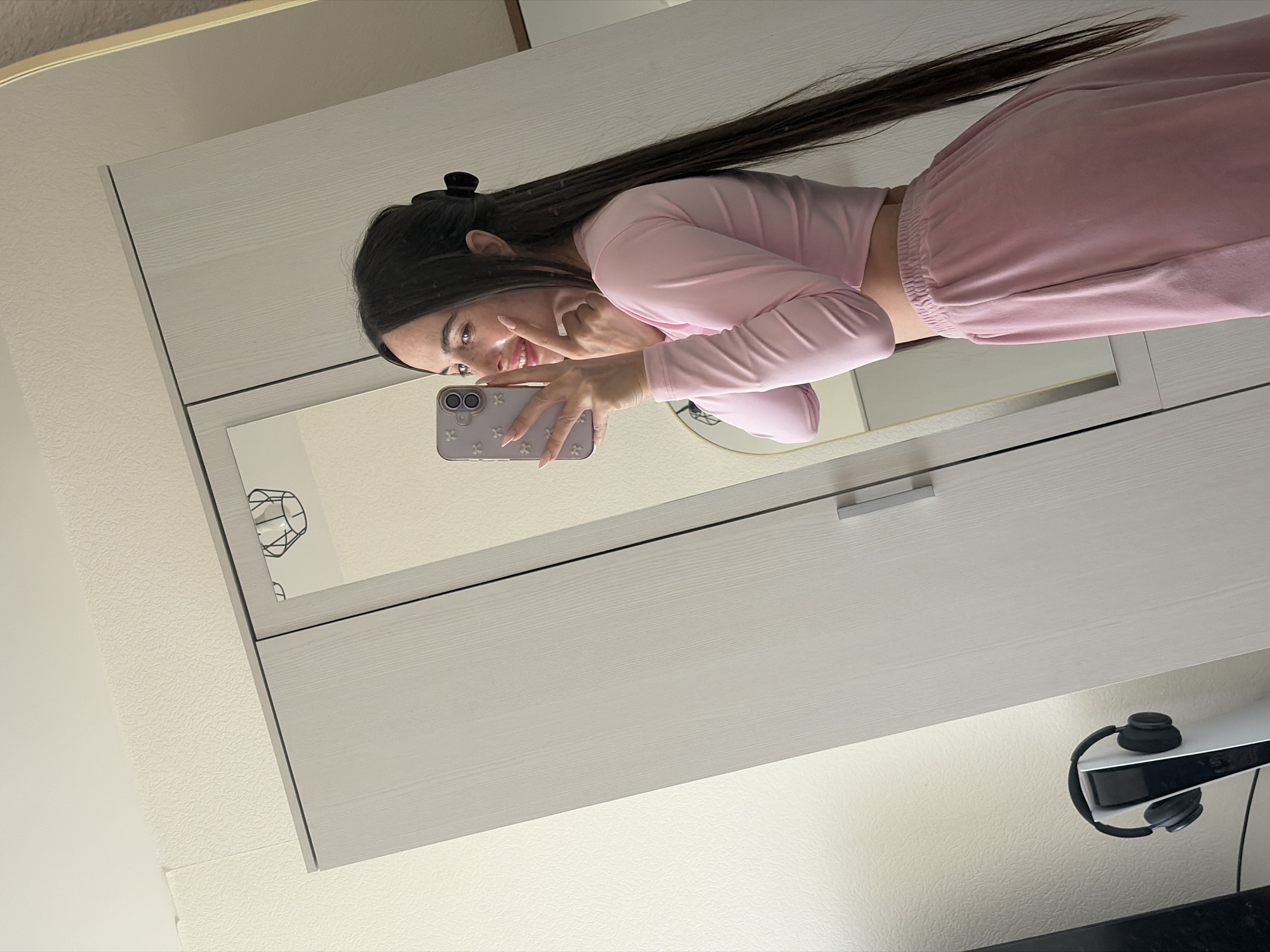 A woman takes a selfie in a mirror inside what appears to be an apartment. She wears a pink top and pants. Her hair is long and dark. In the background is a white cabinet or dresser. There are headphones hanging on the side. The city of Saint Petersburg, Russia, is not discernible from the image.