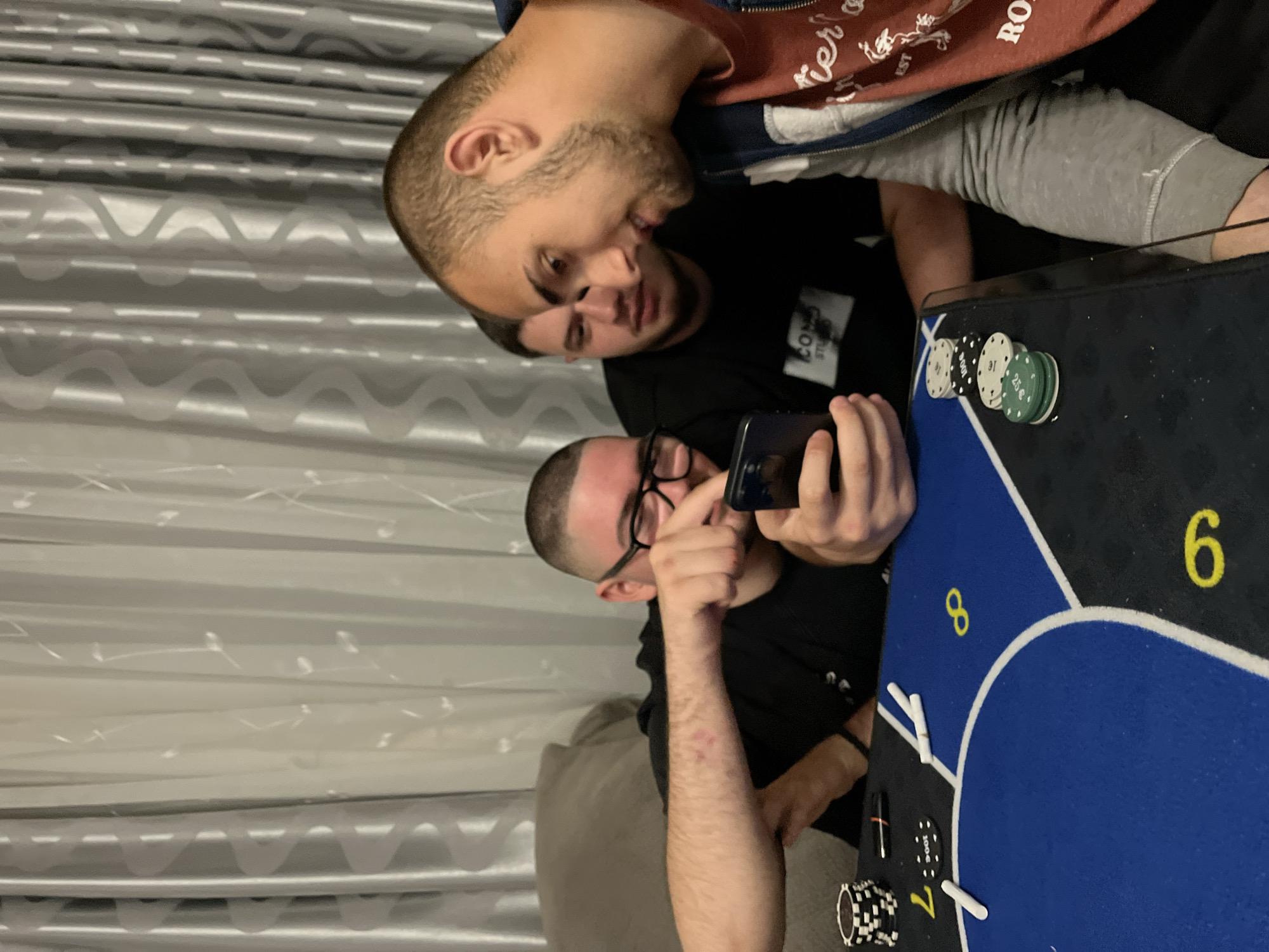 Three men are gathered around a poker table. One man is holding a smartphone, pointing it at something on the table. The table has chips and cards. The background shows a curtained wall. I can't ascertain if this is Burgas, Bulgaria from the image.