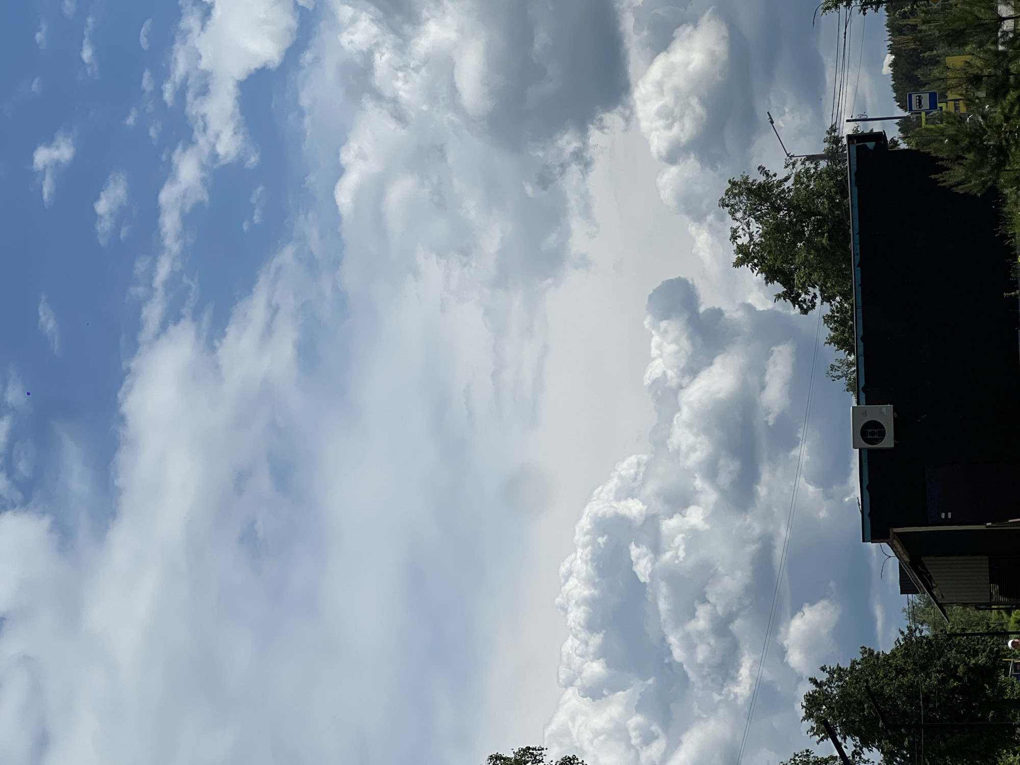 The image shows a daytime sky with scattered white and gray clouds. Part of a dark building is visible on the right side of the frame, with a visible air conditioning unit on its outer wall. Some trees are seen around the building. A bus stop sign is located behind the building. The image seems to be taken in the city of Chaikovsky, Russia.