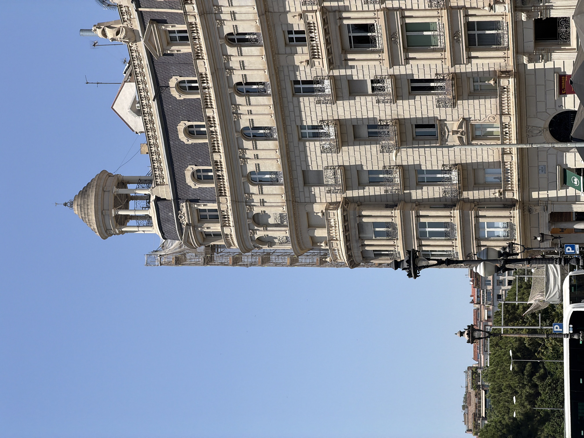 This is an eye-level, medium-shot, exterior photograph of a light-colored, ornate, multi-story building in Barcelona, Spain. The building has a dark, mansard roof with visible shingles and dormer windows. A small, domed tower with white columns and a decorative railing sits atop the building on the left side. The building's facade is detailed with columns, balconies with wrought-iron railings, and multiple arched windows. Scaffolding is visible on the left side of the building, suggesting ongoing construction or renovation.
In the foreground, a white bus is partially visible on the left, and decorative street lamps are present. In the background, a dense line of green trees is visible, with lower-rise buildings with red-tiled roofs behind them. The sky is clear and blue. The overall impression is of a sunny day in a city with historic architecture.